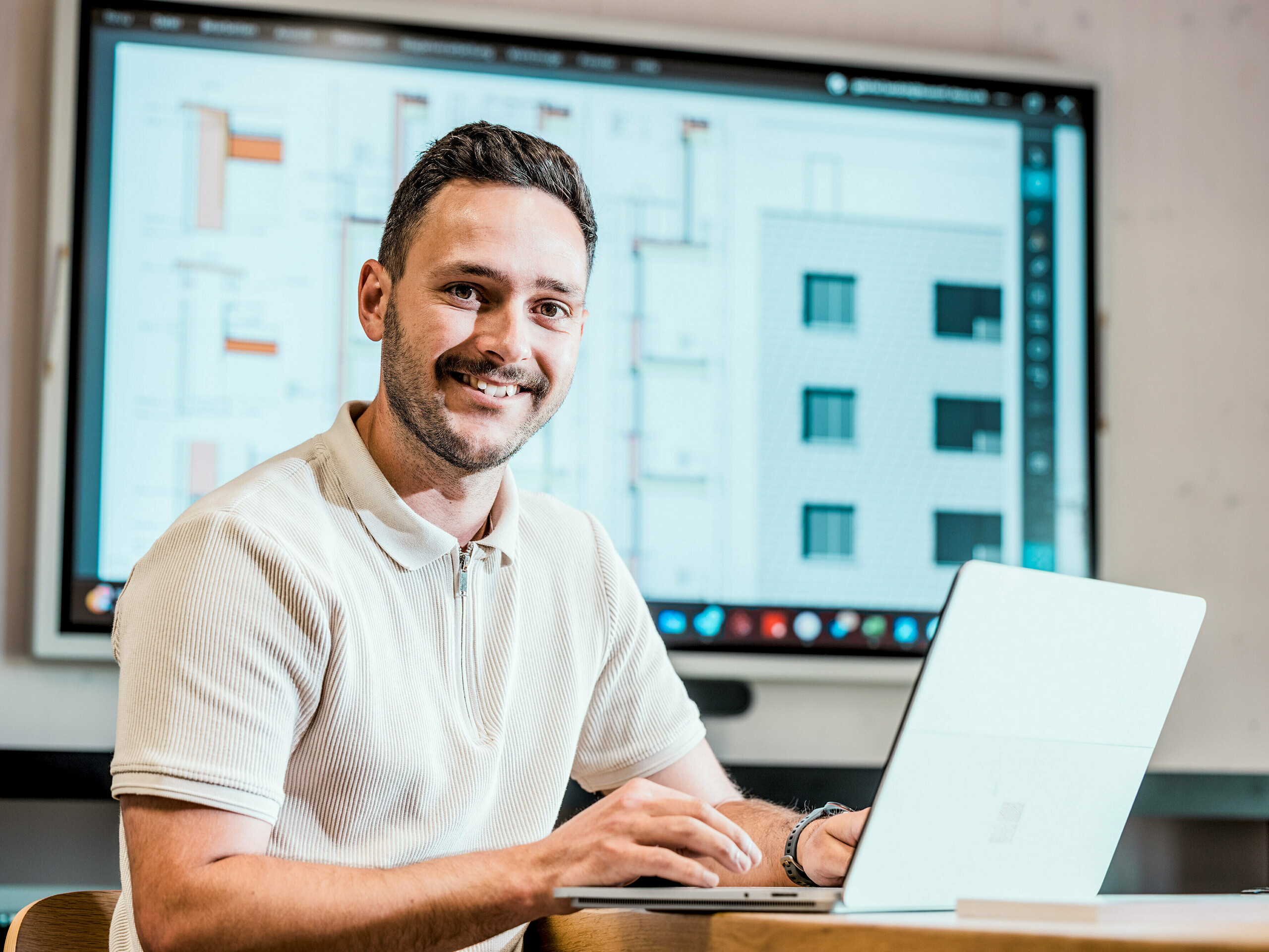 Porträt von Gabriel Landolt, Mitarbeiter der Künzli Davos AG, am Laptop vor einem großen Bildschirm, auf dem digitale Baupläne und Fassadendetails zu sehen sind. Auf dem Monitor sind technische Zeichnungen und Ansichten eines modernen Gebäudes mit schuppenartiger Fassade zu sehen, das mit PREFA Wandschindeln in der Farbe P.10 Hellgrau realisiert wurde. Die innovative Aluminiumfassade überzeugt durch ihr geringes Gewicht, ihre Witterungsbeständigkeit und ihre Langlebigkeit. Eine präzise Planung und die Verwendung hochwertiger PREFA-Produkte sind die Basis für eine formstabile, wartungsarme Gebäudehülle mit architektonischem Anspruch.