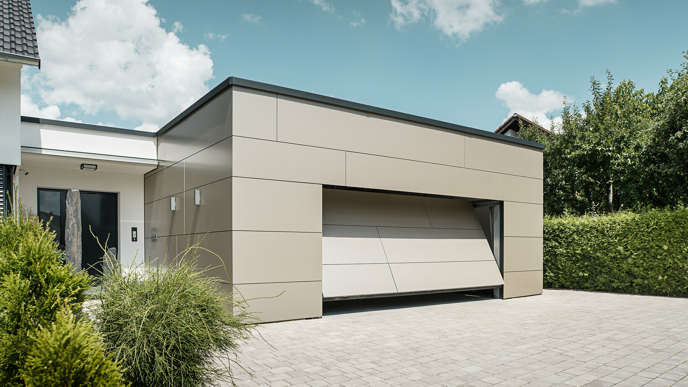 Gesamtansicht einer mit PREFABOND Aluminium Verbundplatten in Bronze verkleideten Garage. Das automatische Garagentor öffnet sich gerade und ist bereits ein Stück geöffnet. Direkt neben der Garage befindet sich der Hauseingang mit anthrazitfarbener Tür und weißer Putzfassade.