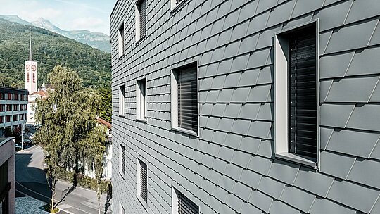 Detailansicht der Fassade des Bernerhauses in Buchs mit präzise verlegten PREFA Wandschindeln in P.10 Hellgrau. Die schuppenartige Aluminiumoberfläche erzeugt ein feines, lebendiges Muster und betont die klar eingeschnittenen Fensteröffnungen mit tiefen Laibungen. Das leichte, witterungsbeständige PREFA Aluminium sorgt für eine homogene, formstabile und wartungsarme Gebäudehülle. Vor der alpinen Kulisse wird die hochwertige Metallfassade zum modernen Eyecatcher mit zeitloser Ausstrahlung.