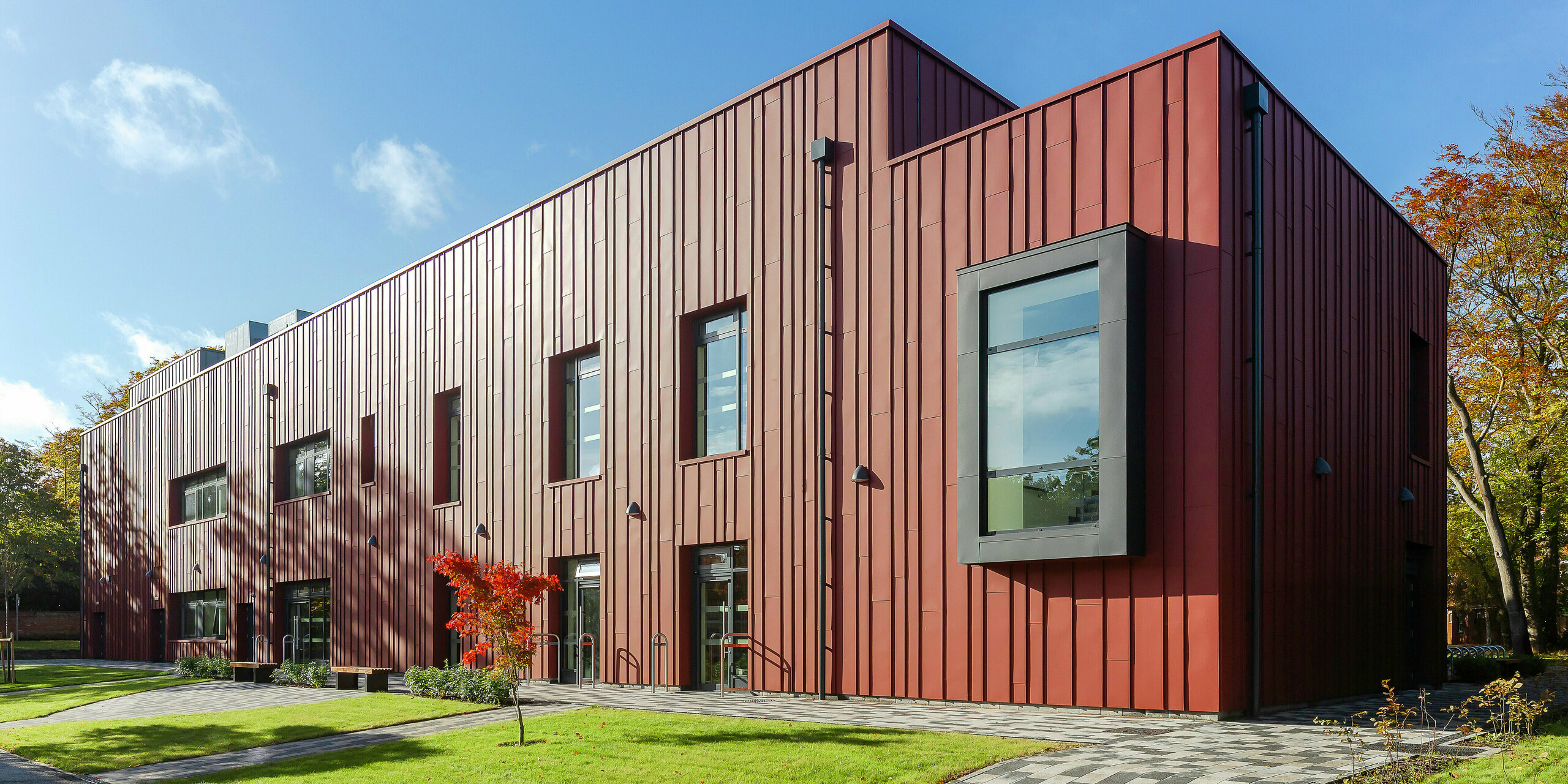 Modern architecture with a robust aluminium façade: the Soldiers' Centre in Pirbright presents an elegant standing seam cladding made of PREFALZ in P.10 oxide red, which blends harmoniously into the green surroundings. The prominent, cantilevered window frames in P.10 anthracite set architectural accents. The paved path and the well-kept green space emphasise the inviting design of the building. Sustainable and aesthetic construction with PREFA aluminium products.