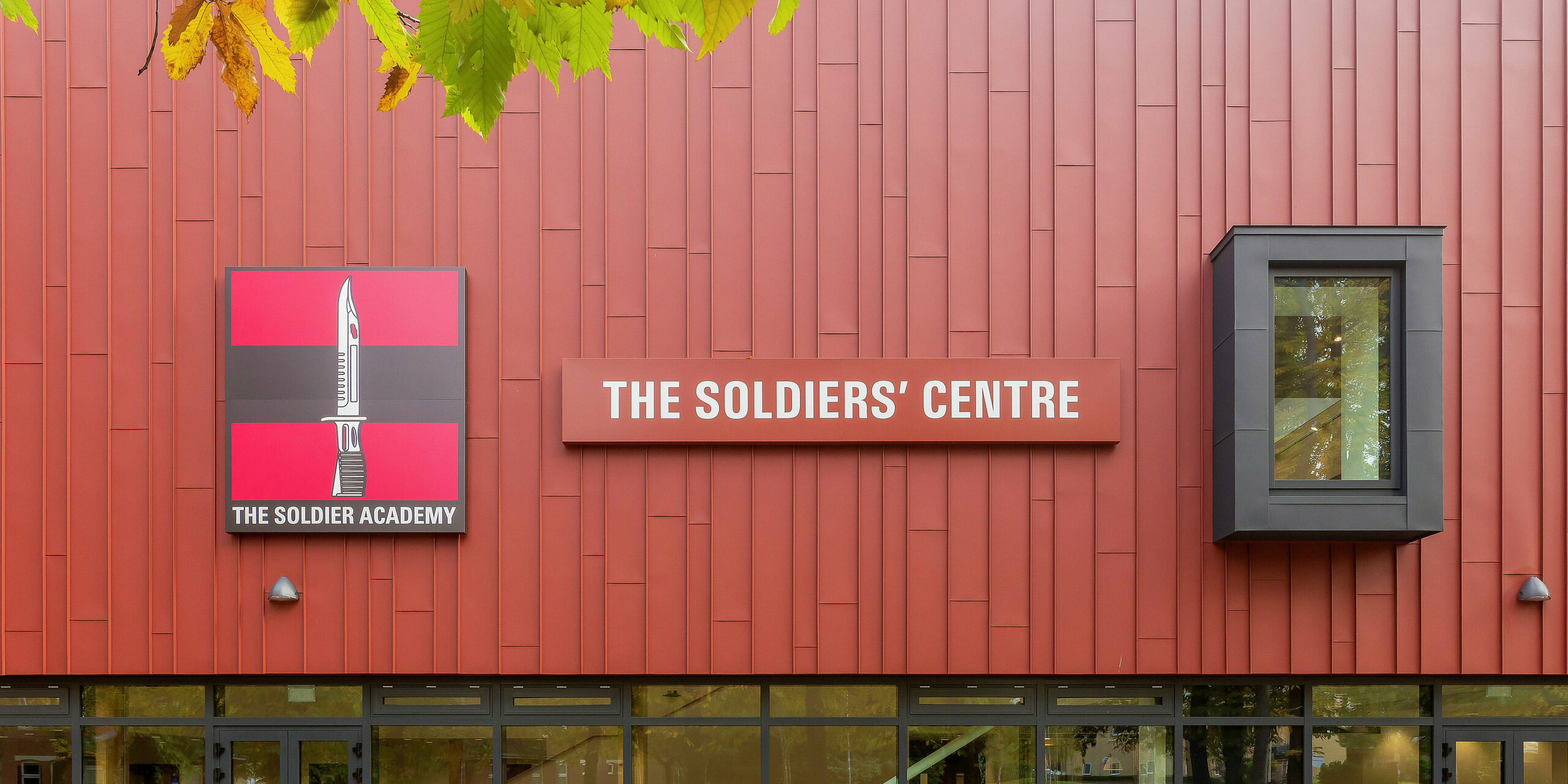 Purpose-built army welfare facility with PREFALZ façade: the precisely manufactured standing seam roof in P.10 oxide red gives the building a striking appearance. The entrance area is impressive with its generous glass fronts, providing brightness and transparency. The 'Soldier Academy' logo and the eye-catching building sign provide a clear visual focus. The window frames in P.10 anthracite round off the design harmoniously. High-quality PREFA aluminium products for durable and aesthetic architecture.