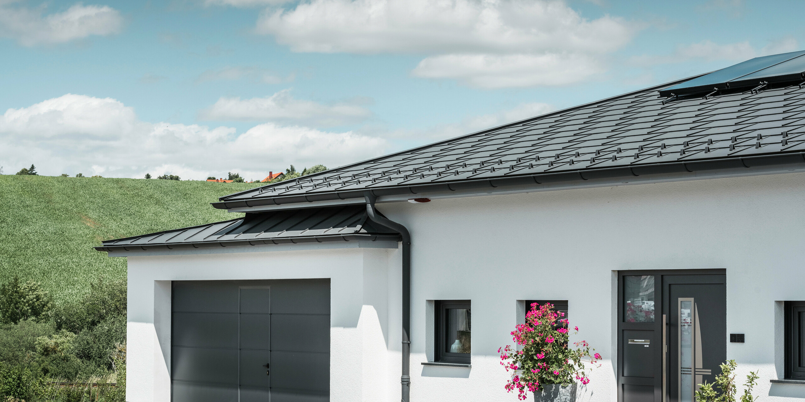 The picture shows the front of a detached house in Ilmenau, Thuringia, with an elegant PREFA roof in P.10 dark grey. The R.16 roof tile gives the house a modern look, while the integrated PREFA roof drainage system with its dark grey half-round gutter and downpipe ensures efficient drainage of rainwater. The garage, fitted with dark grey PREFALZ, blends harmoniously into the overall aesthetic and underlines the quality and style of PREFA aluminium products.