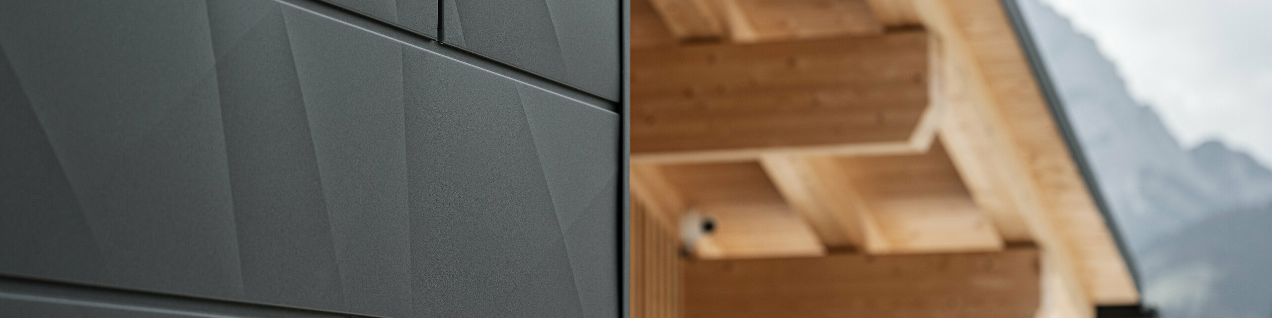 Close-up of the Siding.X façade panels in P.10 black from PREFA on Casa Hoffe in Sappada, Italy. The aluminium façade system has been skilfully installed in a modern, abstract design that forms an attractive contrast to the visible wooden elements of the roof structure. The innovative façade solution blends seamlessly into the alpine ambience and represents the combination of functionality, durability and aesthetic building culture.
