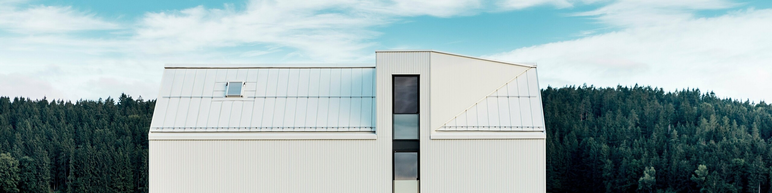 Das Bild zeigt das Einfamilienhaus in Freistadt aus einer Seitenansicht, die die markante, minimalistische Architektur des Gebäudes hervorhebt. Die Fassade aus reinweißem PREFALZ-Aluminium und das steile Satteldach schaffen eine klare, moderne Silhouette, die sich harmonisch in die grüne Umgebung einfügt. Die großen Fensterflächen und das elegante Gaubenfenster betonen das funktionale Design, während das hochwertige Aluminiummaterial sowohl ästhetische Beständigkeit als auch Witterungsresistenz gewährleistet. Das Gebäude strahlt durch seine einfache Form und die Verwendung langlebiger Materialien eine zeitlose Eleganz aus und stellt eine perfekte Kombination aus Architektur und Naturverbundenheit dar.