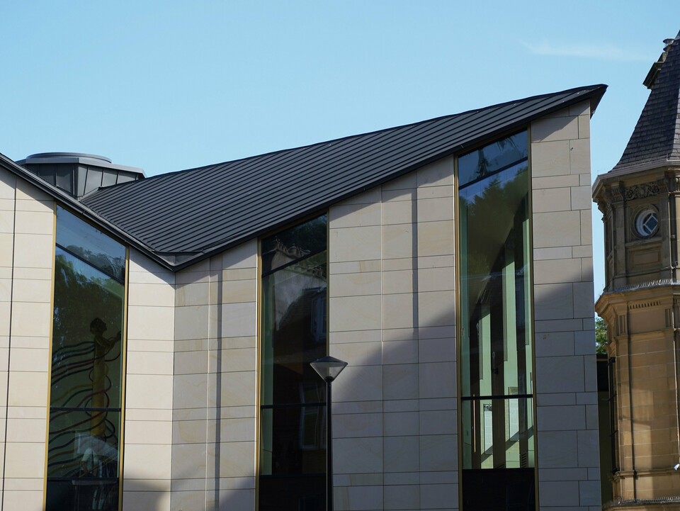 Side view of 'The Great Tapestry of Scotland' museum in Galashiels, which is characterised by a modern PREFALZ roof in P.10 zinc grey. The roof with its clear, geometric lines forms an attractive contrast to the traditional sandstone façades of the neighbouring buildings. The unique roof construction with its striking edges gives the building a contemporary flair and emphasises the combination of historical architecture and modern design.