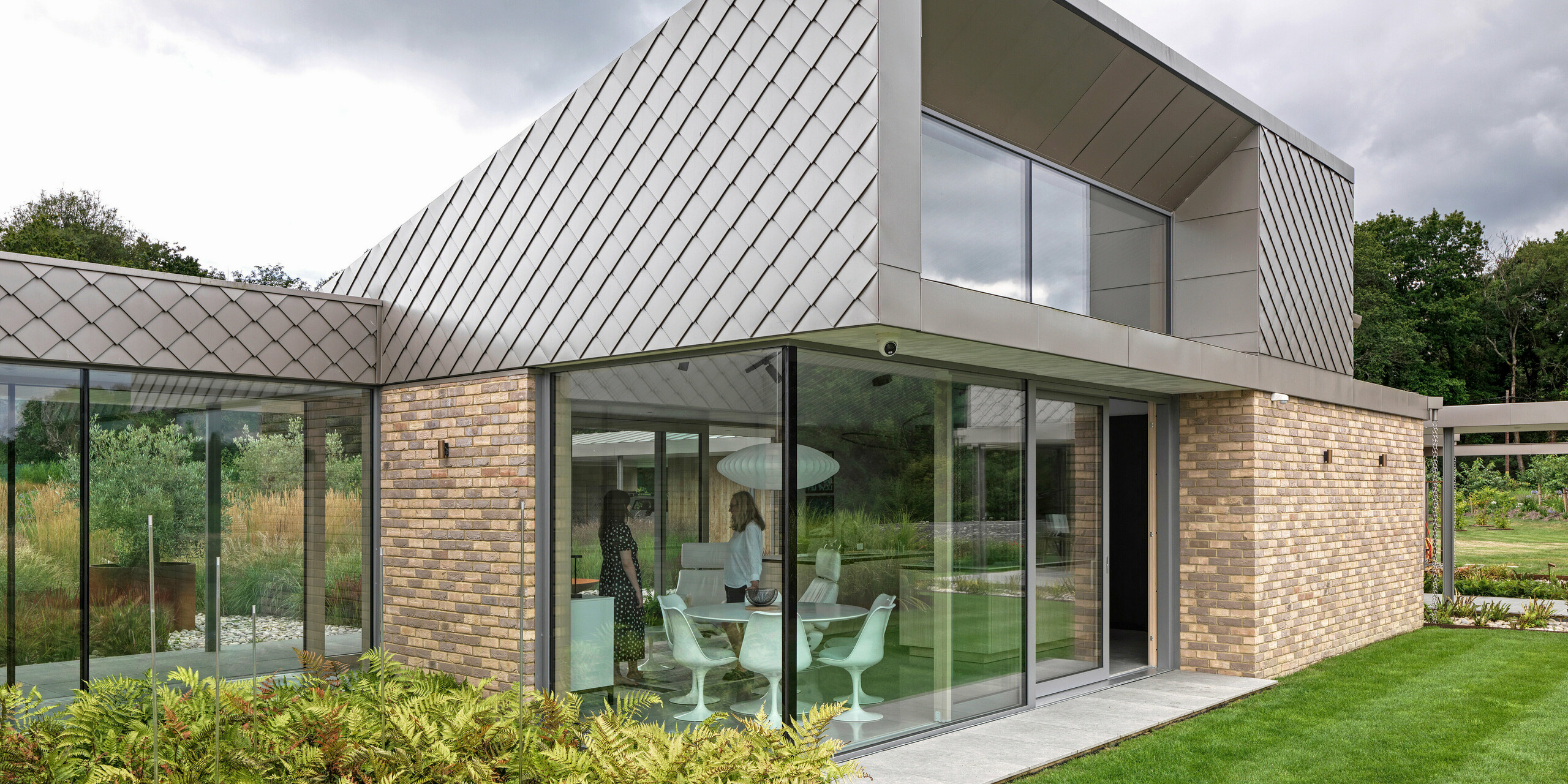 Architecturally striking part of a residential building in West Sussex with a modern façade design featuring PREFA rhomboid façade tiles 20 × 20 in P.10 bronze. The roof surfaces are covered with standing seam and their clear lines emphasise the timeless character of the design. Generous glass fronts create bright living spaces and visually connect the building with the well-kept garden. The high-quality PREFA products offer durability and a uniform appearance with minimal maintenance.