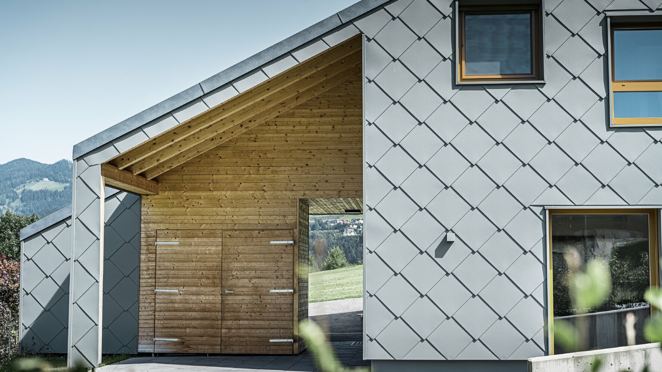 The covered entrance area is made with horizontal wood panelling, the rest of the façade is clad with the large PREFA aluminium rhomboid façade tiles in light grey
