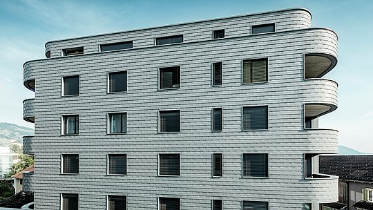 Frontalansicht des Bernerhauses in Buchs mit weich gerundeten Gebäudeecken und durchgehender Fassade aus PREFA Wandschindeln in P.10 Hellgrau. Die schuppenartig verlegte Aluminiumfassade erzeugt ein lebendiges Muster und betont die horizontale Linienführung der geschwungenen Balkonbänder. Präzise integrierte Fensteröffnungen fügen sich harmonisch in die homogene Oberfläche ein. Dank des leichten, witterungsbeständigen und langlebigen PREFA Aluminiums entsteht eine wartungsarme, formstabile Gebäudehülle mit markanter Ausstrahlung vor alpiner Kulisse.