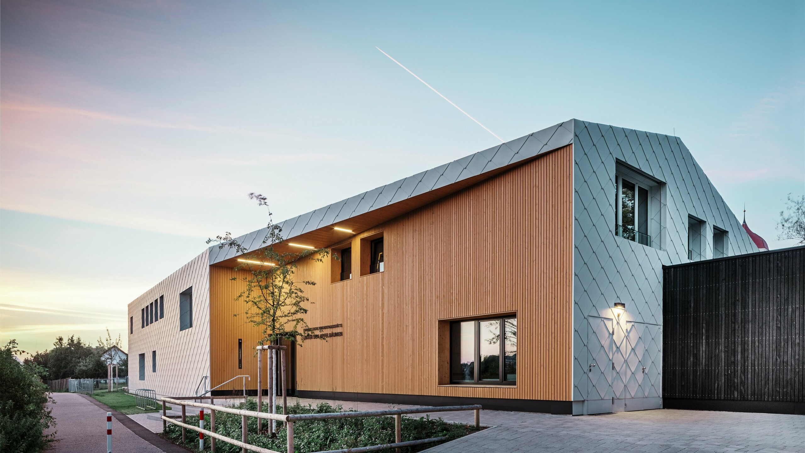A modern nursery school in Ulm at dusk. The roof was designed without eaves. The majority of the façade was clad with large-scale aluminium rhomboids from PREFA in white, while the entrance area was extensively clad with vertical wooden panels.