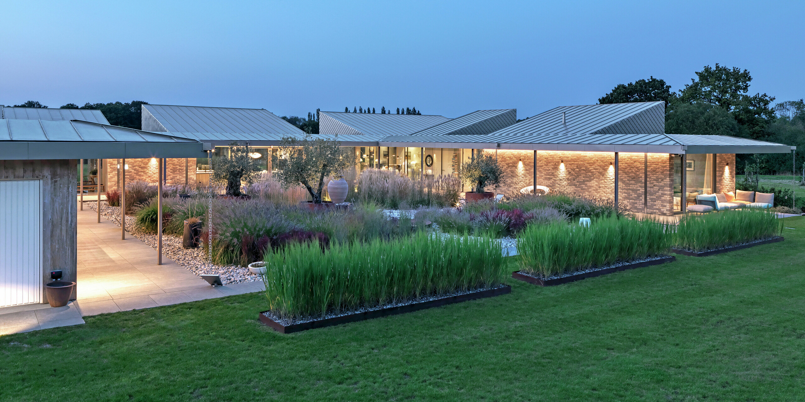 Modern residential building in West Sussex with standing seam roofing and PREFA rhomboid façade tiles 20 × 20 in P.10 bronze – sustainable architecture with durable PREFA roof and façade systems in harmony with nature. The photo was taken at dusk, immediately highlighting the high-quality lighting. At the centre of the property is an impressive garden with spectacular plants.