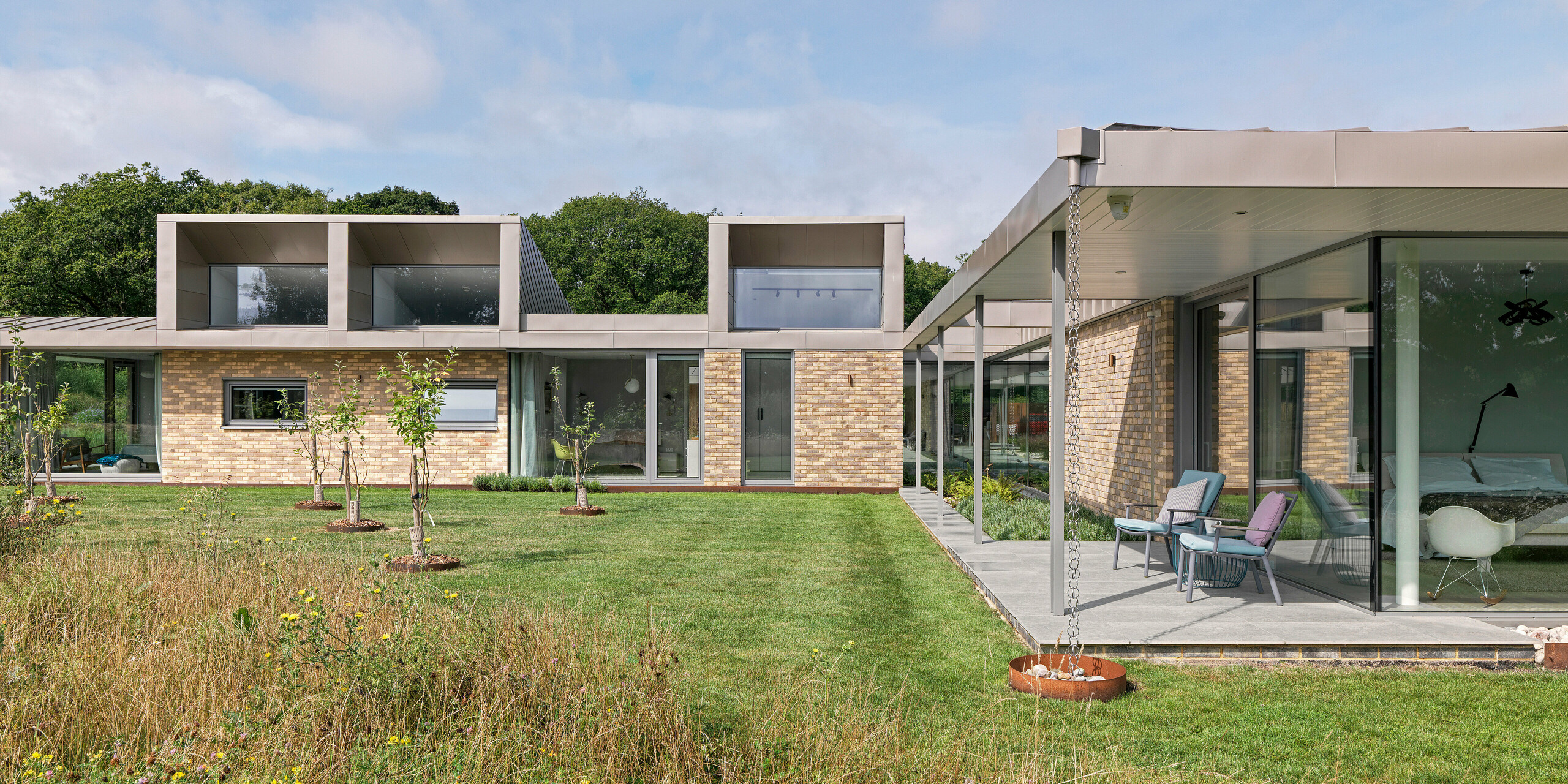 Side view of a luxurious residential building in West Sussex with striking upper floor structures clad with PREFA rhomboid façade tiles 20 × 20 in P.10 bronze. The flat roof is made of standing seam, also in P.10 bronze, and emphasises the minimalist design. Large glass façades, a covered terrace with filigree supports and rainwater drainage chains round off the sustainable architectural concept with durable and maintenance-free PREFA aluminium products.