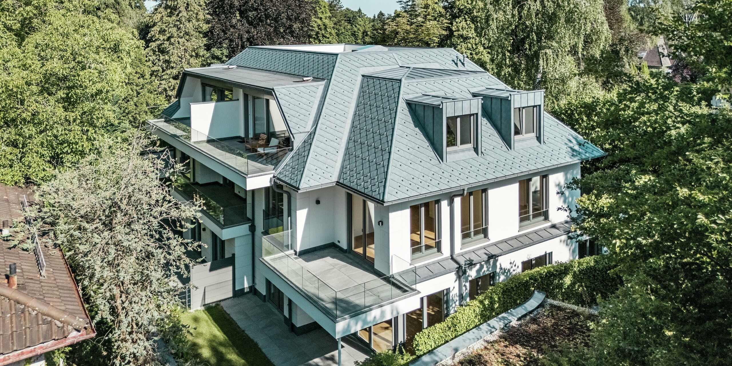 Luftaufnahme eines modernen Mehrfamilienhauses mit auffälliger Dachgestaltung, gedeckt mit PREFA Dachrauten 29 × 29 in der eleganten Farbe P.10 Steingrau. Das imposante Mansard-Flachdach in Kombination mit stilvollen Flachdachgauben aus PREFALZ bildet einen markanten Blickfang. Die hochwertigen Aluminiumprodukte fügen sich perfekt in die grüne Umgebung ein und bieten sowohl ästhetische als auch funktionale Vorteile. Die präzise Verarbeitung und Witterungsbeständigkeit der PREFA-Produkte garantieren Langlebigkeit und schützen das Gebäude vor den Elementen.