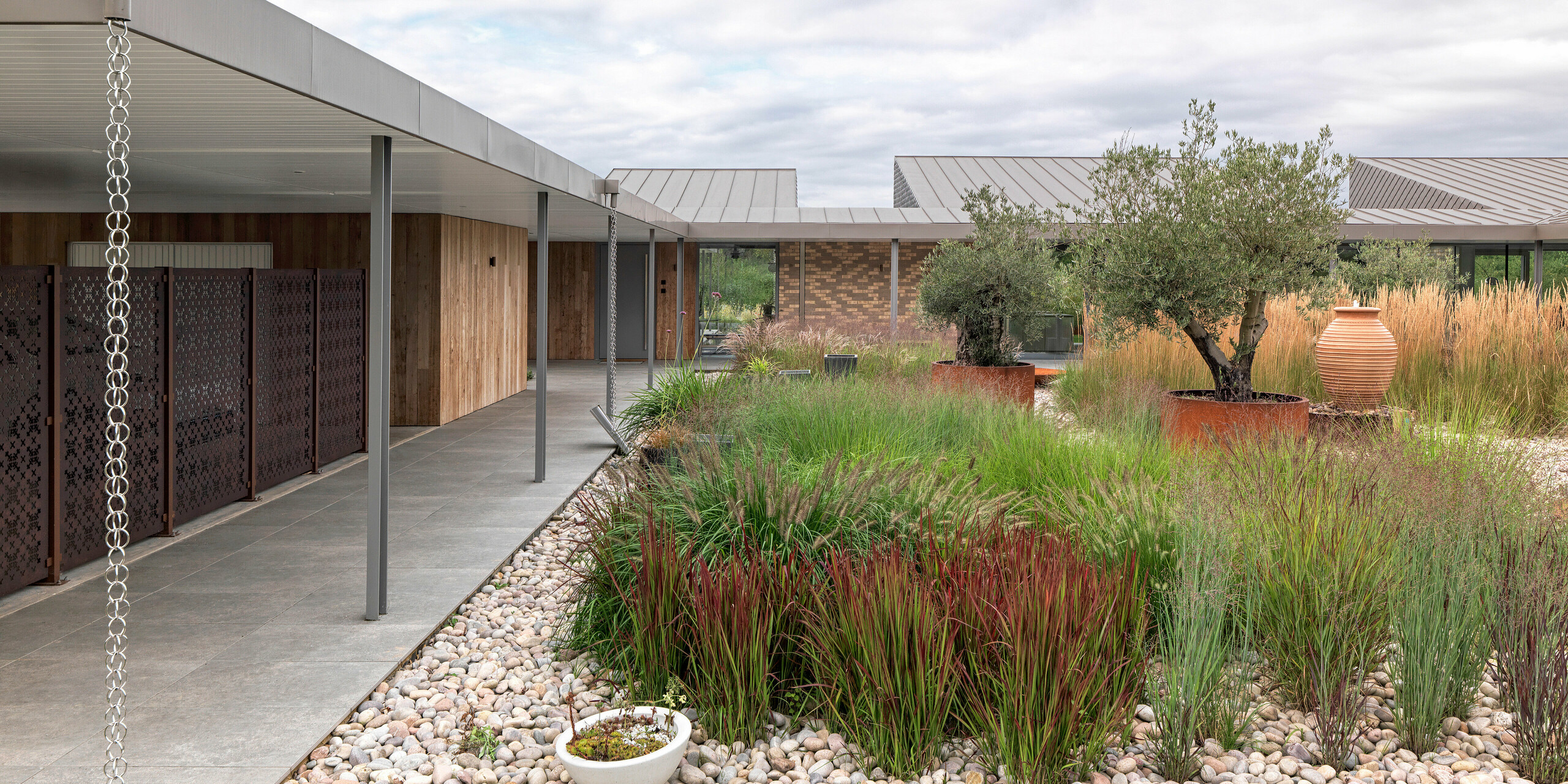 Detailed view of the modern residential building in West Sussex with a flat roof made of standing seam in P.10 bronze. The covered, clearly structured terrace area with filigree columns, stylishly integrated rainwater drainage chains and a varied garden design with ornamental grasses, olive trees and natural stones can be seen. The durable PREFA roof products made of aluminium emphasise the modern lines and blend harmoniously into the sustainable architectural concept.