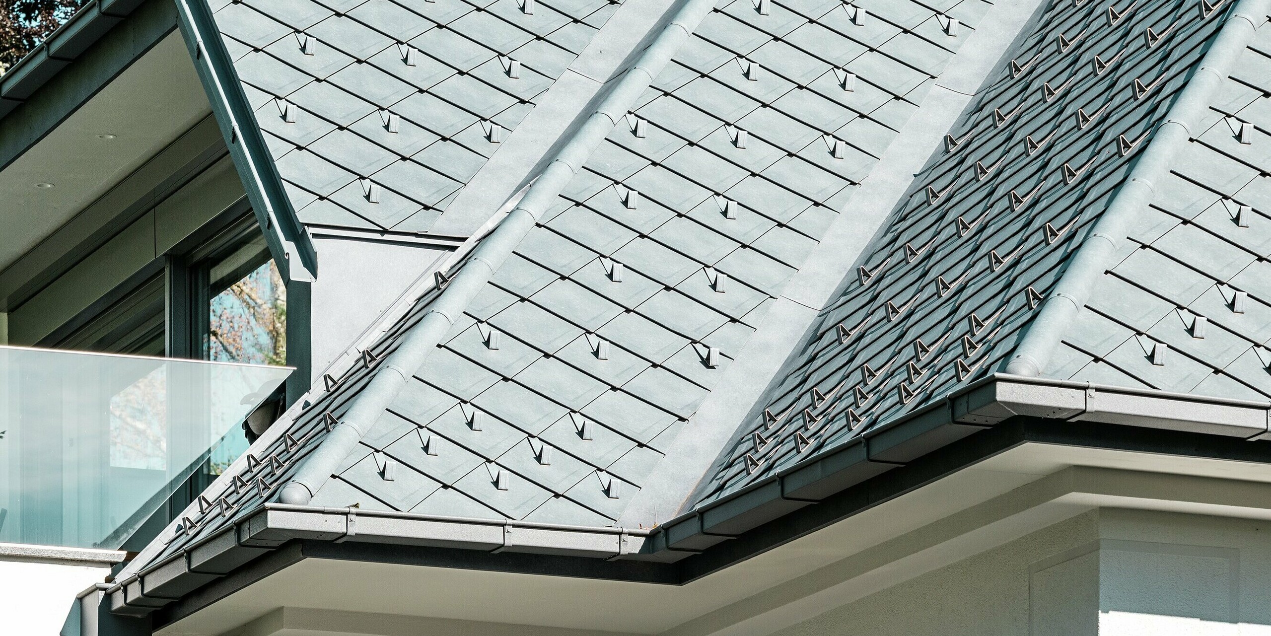 Nahaufnahme eines modernen Mehrfamilienhauses mit einem stilvollen Dach aus PREFA Dachrauten 29 × 29 in der Farbe P.10 Steingrau. Die Dachflächen werden durch integrierte Schneestopper ergänzt, die für zusätzliche Sicherheit im Winter sorgen. Die formschöne PREFA Dachentwässerung mit Kastenrinne und Ablaufrohr rundet das Design ab und garantiert effizienten Wasserabfluss. Die Kombination aus hochwertigen Materialien und durchdachtem Design macht das Gebäude nicht nur ästhetisch ansprechend, sondern auch funktional und langlebig.