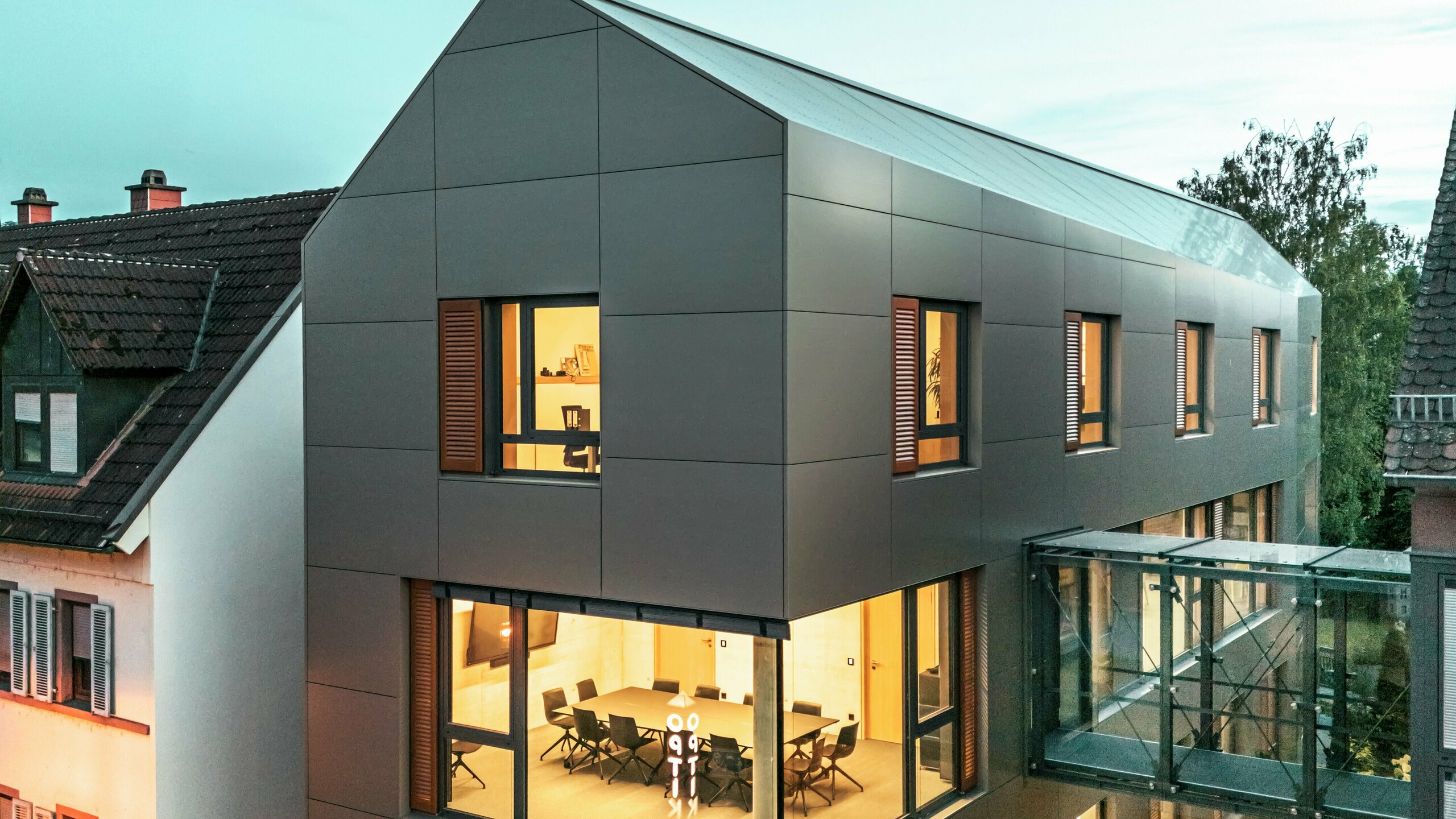 The image shows the town hall in Alzenau at dusk, with a modern façade made of aluminium composite panels from PREFA in an elegant bronze tone. The glass connecting passageway, which links the building to an adjacent section, stands out visually. The large windows offer views of the interior, which is illuminated by warm light. The clear geometric shape of the façade and the surrounding green space reinforce the modern, contemporary look of the building.