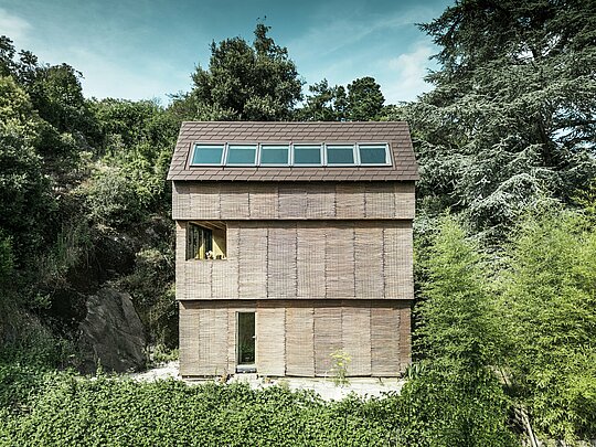 Moderne Architektur in Frankreich – ein Haus mit PREFA-Dachschindeln in der Farbe P.10 Walnussbraun und einer Fassade aus Weidenholz, gelegen an einem steilen Hang mit großzügigen Oberlichtern.