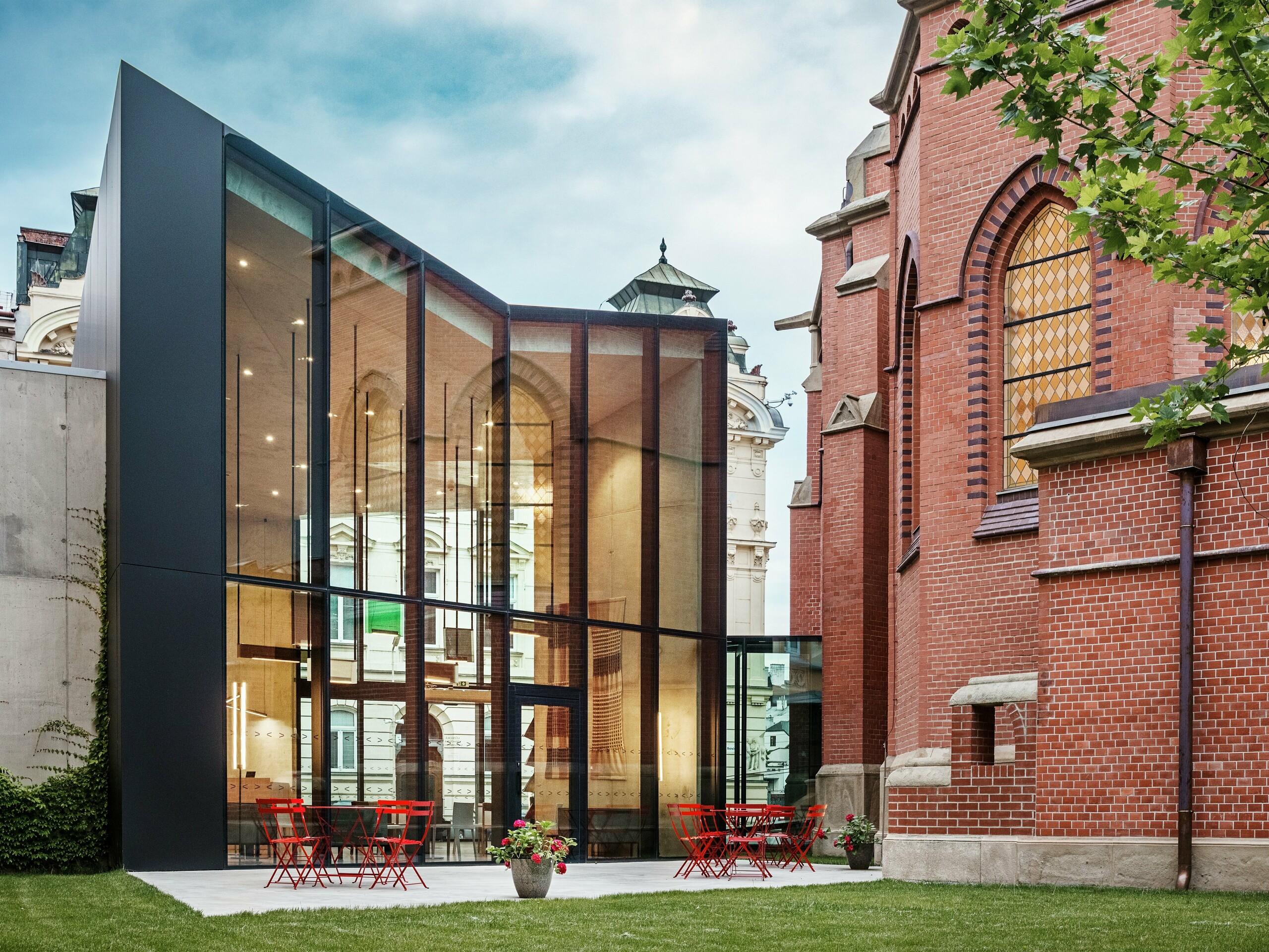 Das Bild zeigt den faszinierenden Neubau in Olmütz, der die historische Rote Kirche mit einer modernen Bibliothek verbindet. Die markante schwarze Fassade aus PREFABOND Aluminium-Verbundplatten setzt einen starken Kontrast zu den warmen, rotgebrannten Backsteinen der Kirche. Große Glasflächen bieten einen klaren Blick auf das Innere des Gebäudes, wo sich eine moderne, lichtdurchflutete Atmosphäre entfaltet. Auf der Terrasse laden rote Tische und Stühle zum Verweilen ein und schaffen eine harmonische Verbindung zwischen dem historischen Bauwerk und der zeitgenössischen Architektur. Diese elegante Integration von Alt und Neu zeigt die Vorteile der PREFABOND Platten – sie sorgen nicht nur für eine moderne, ästhetische Wirkung, sondern bieten auch herausragende Langlebigkeit und Funktionalität.