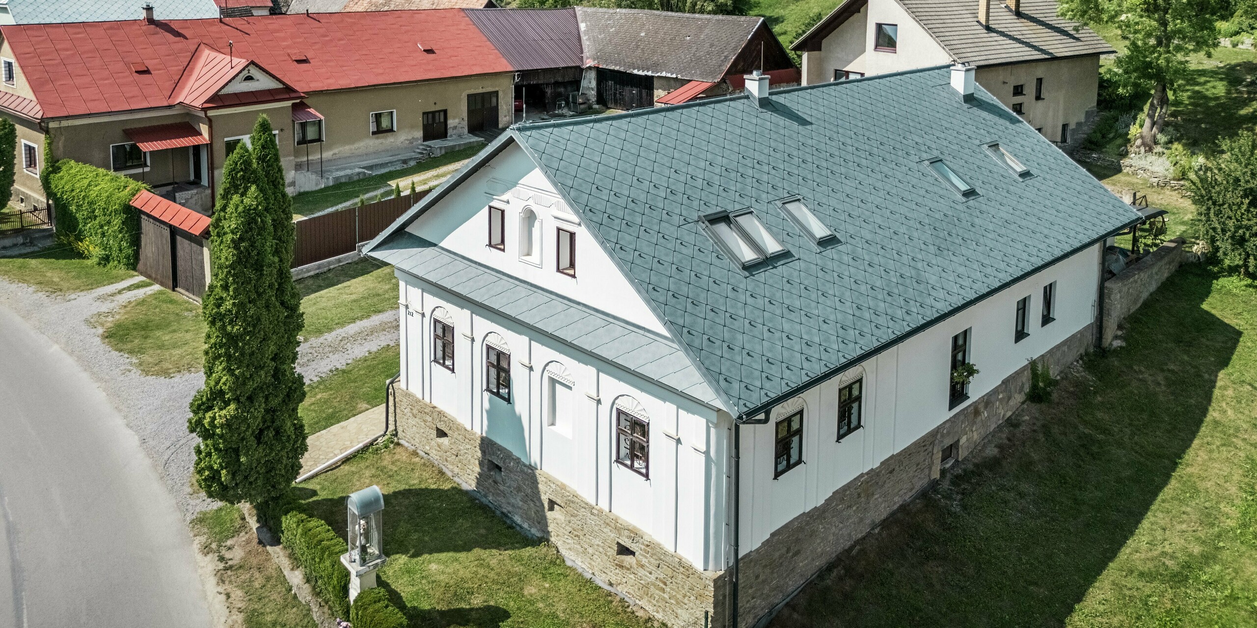 Das Bild zeigt das sanierte Handwerkerhaus in Klin, Slowakei, mit einem Satteldach eingedeckt mit PREFA Dachrauten 44 × 44 in P.10 Steingrau. Die symmetrische Dachstruktur mit integrierten Dachfenstern bietet nicht nur eine ästhetische Verbesserung, sondern sorgt auch für viel natürliches Licht im Inneren des Hauses. Das Aluminiumdach ergänzt perfekt die klassische Fassade mit Natursteinmauerwerk und bietet eine hervorragende Langlebigkeit, Korrosionsbeständigkeit und Leichtigkeit. Diese nachhaltige Dachlösung ist ideal für die Sanierung historischer Gebäude und garantiert gleichzeitig optimale Funktionalität und Schutz vor Witterungseinflüssen.