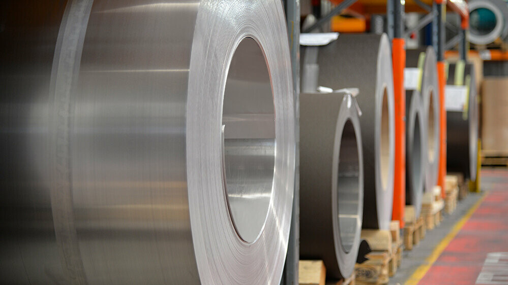 Aluminum coils stored on wooden pallets, with a plain aluminum coil in the foreground and further aluminum coils and sheets behind it.