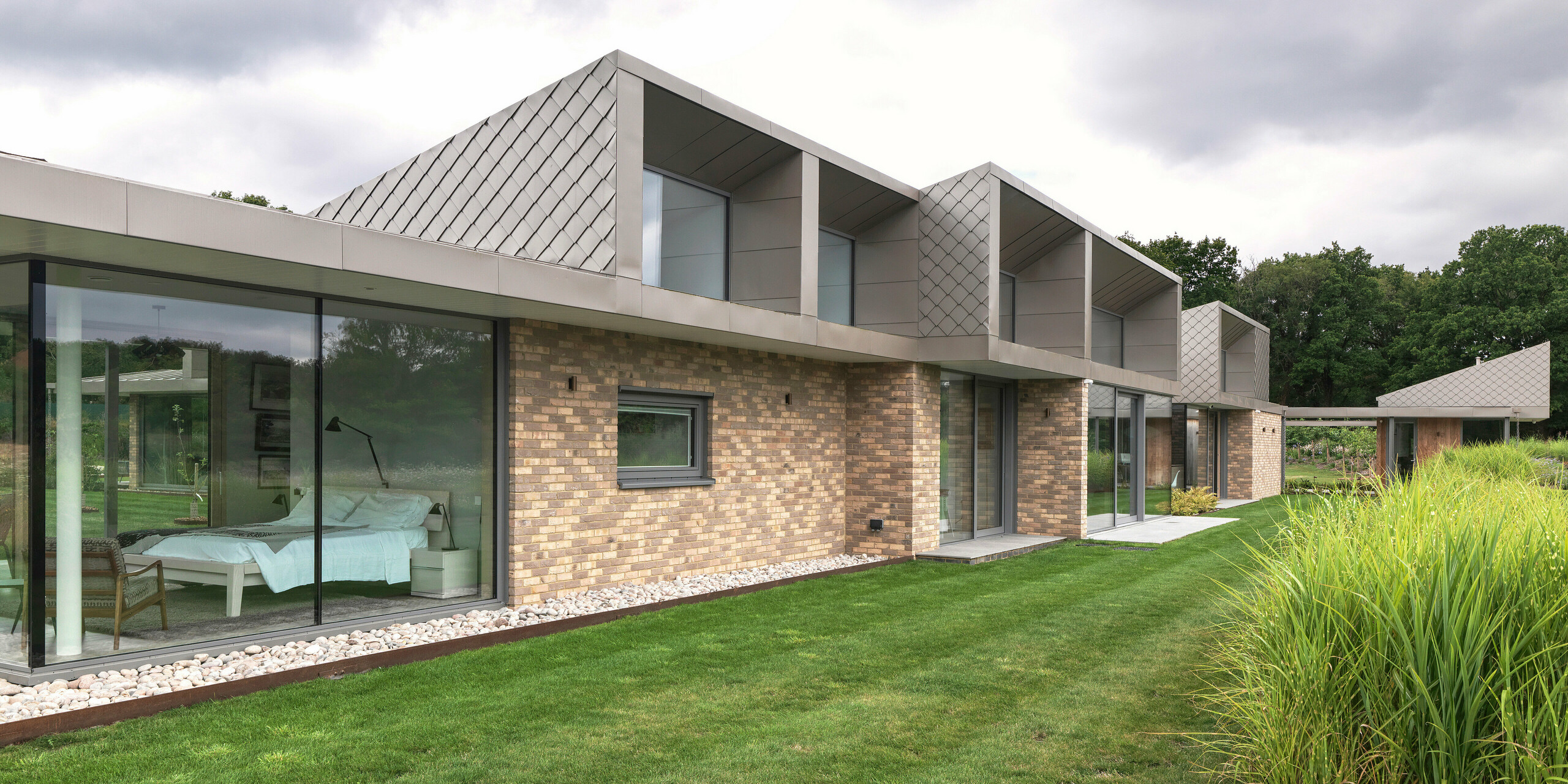 Side view of the modern residential building in West Sussex with striking superstructures on the upper floor, clad with PREFA rhomboid façade tiles 20 × 20 in P.10 bronze.The flat-sloping aluminium roof forms an elegant and weather-resistant junction. Large glazed areas on the ground floor seamlessly connect the interior and exterior spaces, giving the building a high-quality, timeless appearance.