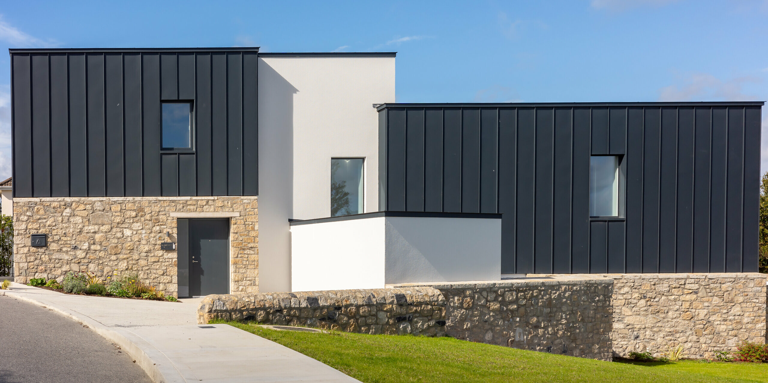 Modern residential building in Watson Place with clean lines, natural stone walls and striking PREFALZ standing seam roof and façade cladding in P.10 anthracite. The smooth aluminium surfaces create an elegant contrast to the stone and plaster and offer long-lasting protection from the weather. The interplay of materials gives the building a high-quality, timeless appearance.