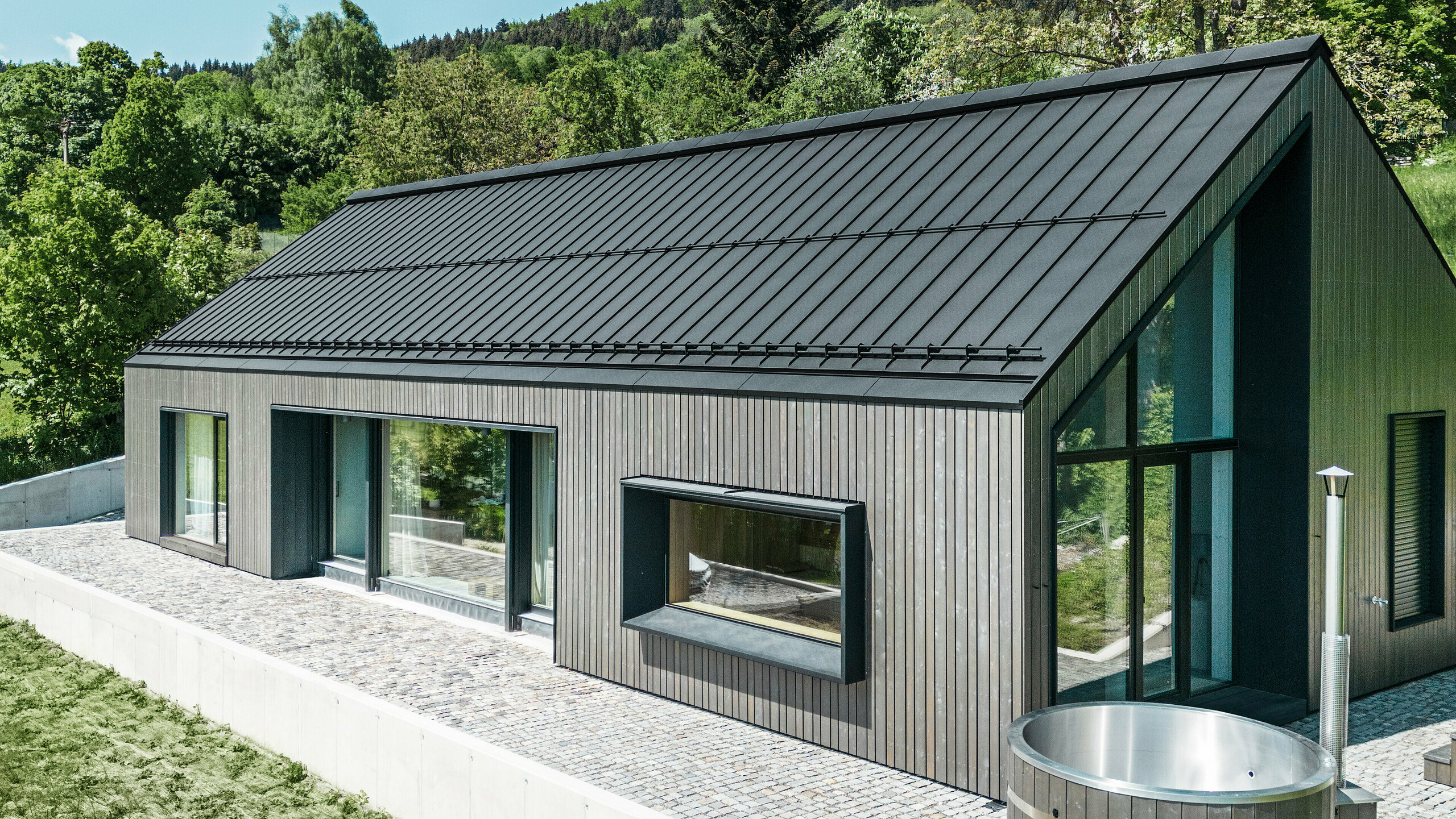 A modern wooden house in Kuncice (CZ) was equipped with large windows. The dark wooden house has a PREFALZ (standing seam) roof system in the colour P.10 black. In the rear outdoor area, there is a hot tub with wooden panelling.