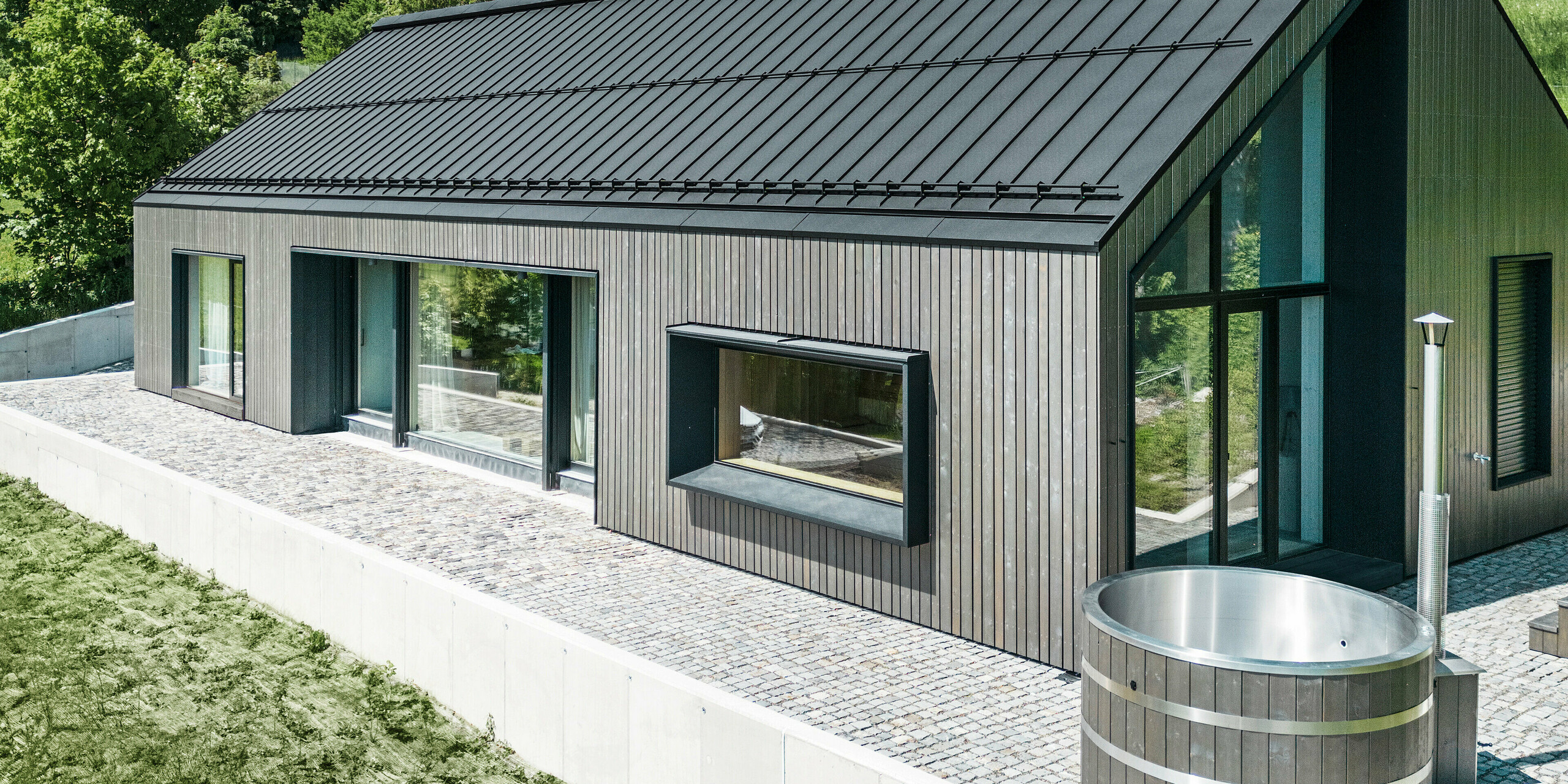 A modern wooden house in Kuncice (CZ) was equipped with large windows. The dark wooden house has a PREFALZ (standing seam) roof system in the colour P.10 black. In the rear outdoor area, there is a hot tub with wooden panelling.