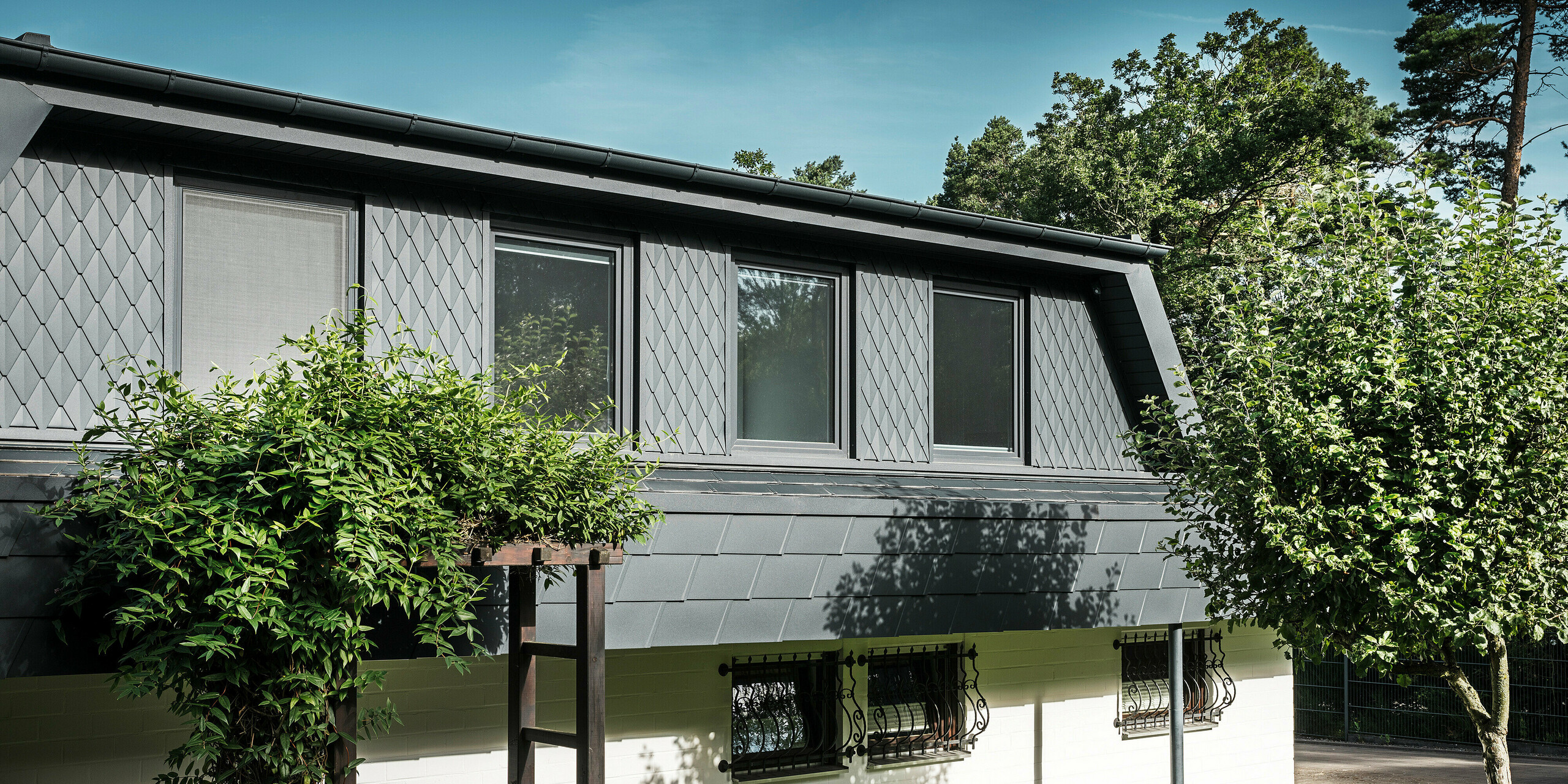 Seitenansicht eines Einfamilienhauses in Mechernich mit großzügigen Trapezgauben. Die Gauben sind mit kleinen PREFA Dachrauten in P.10 Anthrazit versehen, während das Dach mit den bewährten PREFA Dachschindeln DS.19 ebenfalls in P.10 Anthrazit eingedeckt ist. Die Aluminium-Dachrauten und -schindeln sorgen für eine wetterbeständige, ästhetische und nachhaltige Lösung für Dach und Fassade. Das traditionelle Einfamilienhaus ist von einem gepflegten Garten und zahlreichen Bäumen umgeben.