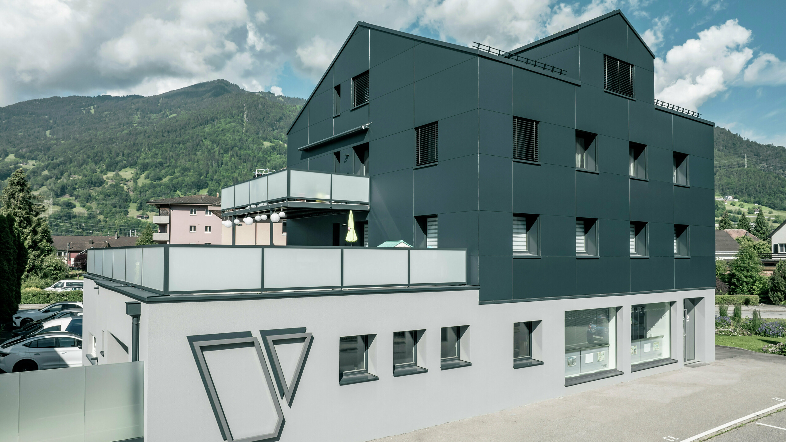 Das Architekturbüro Voser in Landquart, im Schweizer Graubünden, mit einer PREFA Aluminiumfassade in Anthrazit. Das Gebäude wurde saniert und mit PREFABOND Aluminium Verbundplatten ausgestattet. Die Gestaltung kombiniert klare Linien und hochwertige Materialien. Auffällig sind die großen Balkone mit Glasbrüstungen und das prägnante Logo des Architekturbüros Voser an der Gebäudeseite. Im Hintergrund ist eine grüne Berglandschaft zu erkennen.