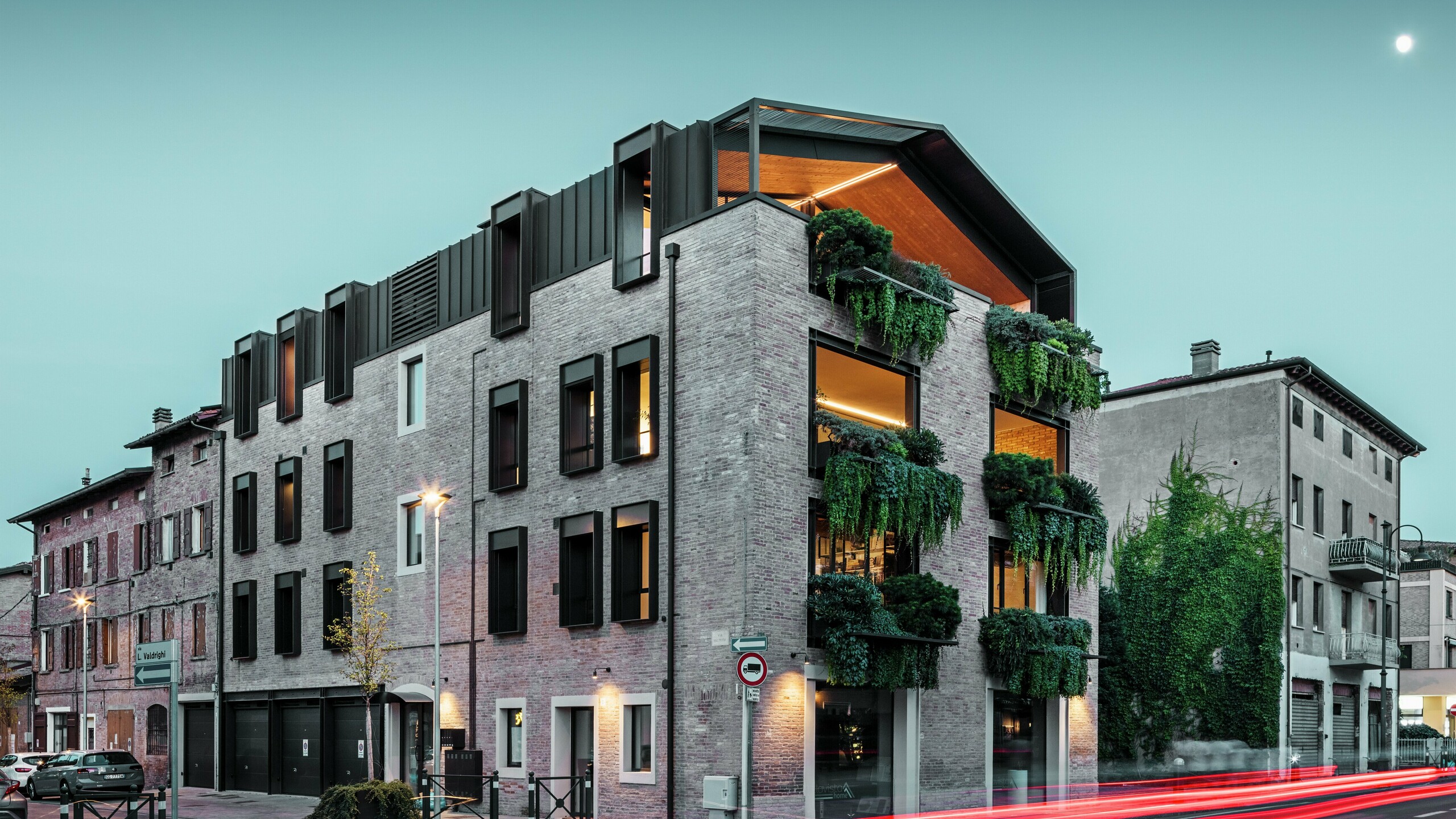 Nachhaltiges Wohn- und Geschäftshaus in Italien mit PREFALZ Aluminiumdach in P.10 Nussbraun, begrünter Backsteinfassade und moderner Architektur bei Abendstimmung mit leuchtenden Straßenlaternen.