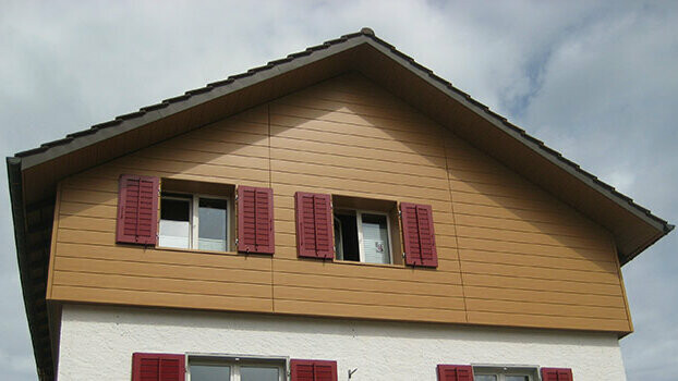 Gable cladding of a traditional house with a gable roof. The gable is clad with PREFA natural oak sidings, installed horizontally. The windows have red shutters.