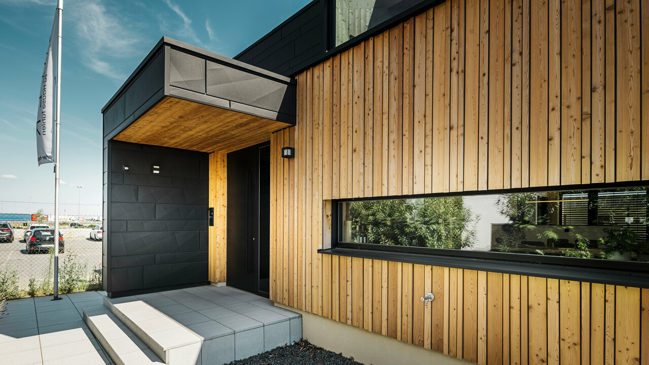Side entrance of the model house at the prefabricated house centre Blaue Lagune in Vösendorf, with a modern façade made of natural wood and black PREFA Siding.X panels. The picture shows the elegant combination of wood cladding and aluminium in the high-quality P.10 black coating.