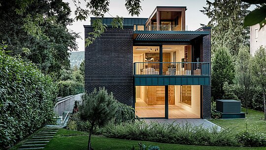 Die Rückseite der modernen Mehrgenerationen‑Villa in Budapest ist eingebettet in eine üppig begrünte Gartenlandschaft. Große Glasflächen öffnen den Wohnbereich nach außen und lassen warmes Innenlicht auf die strukturierte Klinkerfassade treffen. Die klaren Linien des Gebäudes werden durch PREFALZ Aluminium in P.10 Anthrazit betont, das als Balkonverkleidung und Fassadenelement langlebige Akzente setzt. Die Kombination aus Backstein, Aluminium und viel Grün unterstreicht den zeitlosen, hochwertigen und architektonisch anspruchsvollen Charakter des Hauses.