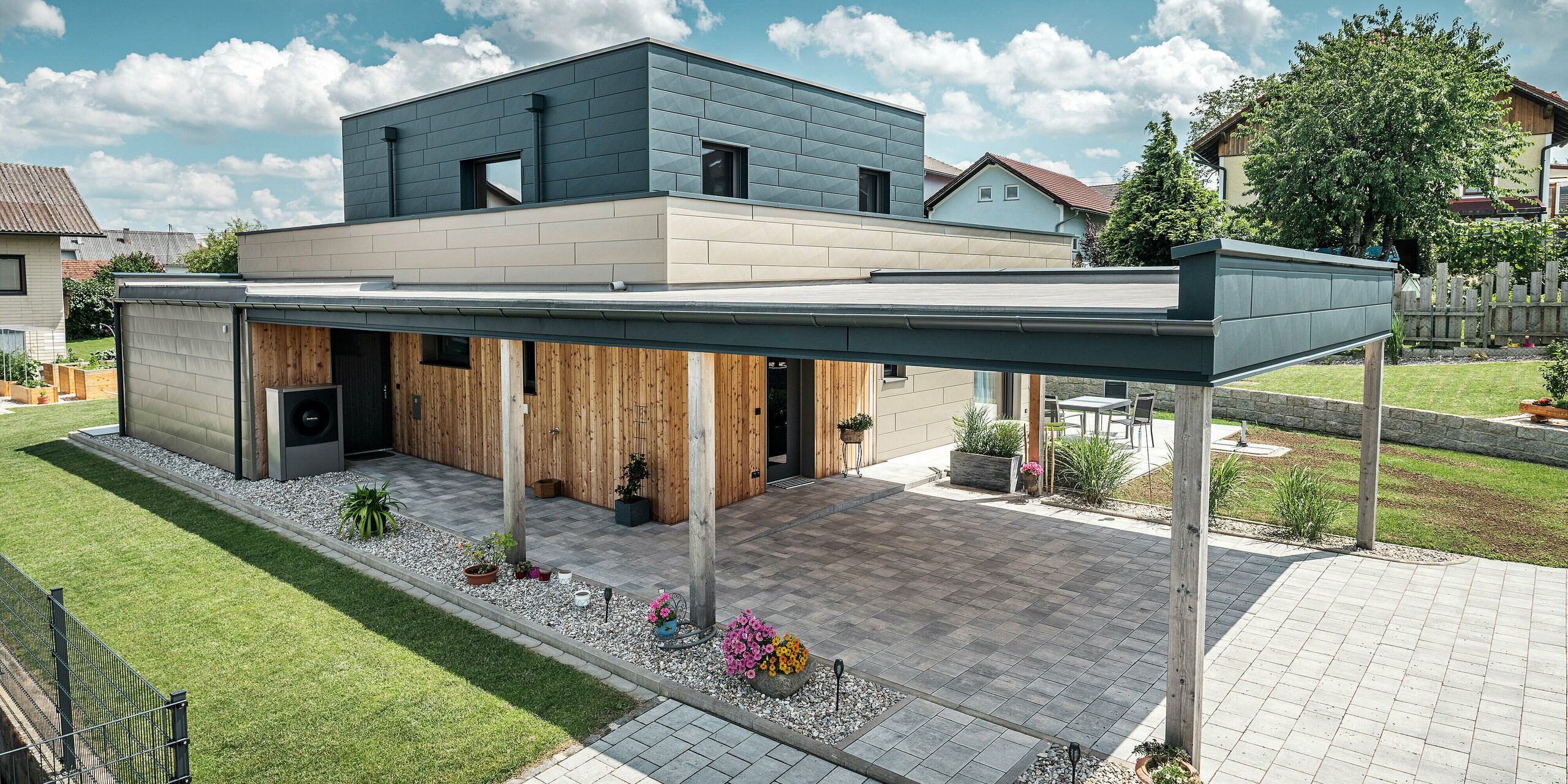 Aerial view of the driveway of a modern detached house in Wippenham, Austria, with a two-tone PREFA aluminium façade in bronze and anthracite. The house showcases the stylish integration of Siding.X façade panels and wood cladding. The covered carport is clearly visible and seamlessly integrated into the architecture of the house. The well-kept garden and terrace area emphasise the modern and functional design of the building.