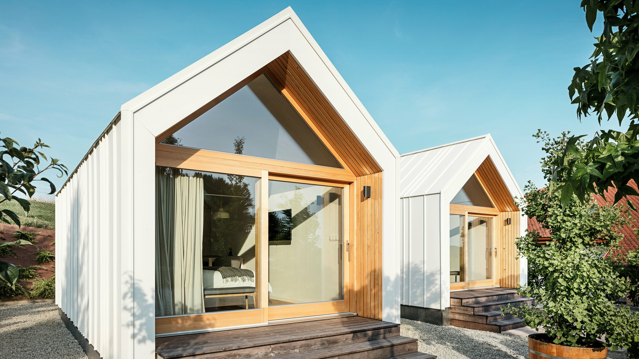 Front view of the two tiny houses of the 'Pri Momi' guest accommodation in Kančevci, Slovenia, with large glass fronts, designed with PREFALZ roof and façade cladding in P.10 pure white and natural wood. The minimalist micro houses have wide wooden steps leading up to the entrances and offer a direct view into the cosy interiors. The clear lines and the combination of aluminium and wood underline the modern and inviting design of the holiday accommodation.