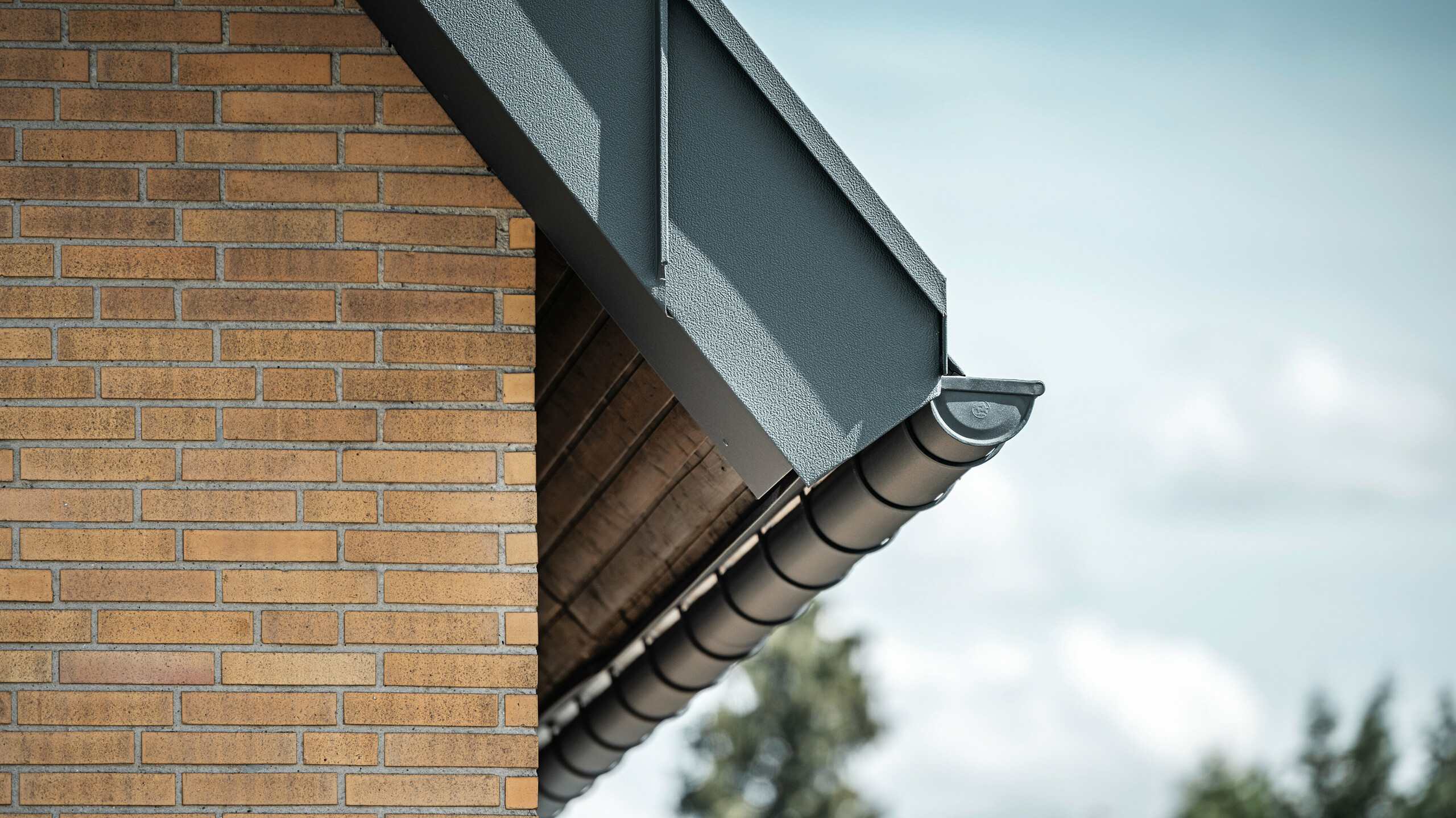 Detailed view of an anthracite-coloured roof drainage system from PREFA. The half-round gutter stands out clearly against the warm tones of the brick wall and emphasises the precise workmanship and quality of the materials used.