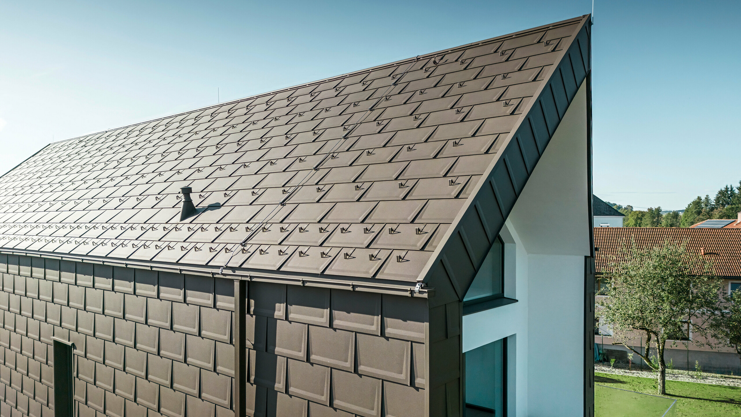 A perspective view of the roof and façade of a modern detached house in Neukirchen, Austria. The PREFA R.16 roof tile in brown P.10 provides an aesthetic and functional roof covering that blends perfectly into the architectural design of the house. The sharp ridge of the roof and the precise arrangement of the roof tiles demonstrate the quality of PREFA. The small format aluminium products blend perfectly into the white plastered façade and the surrounding green landscape.