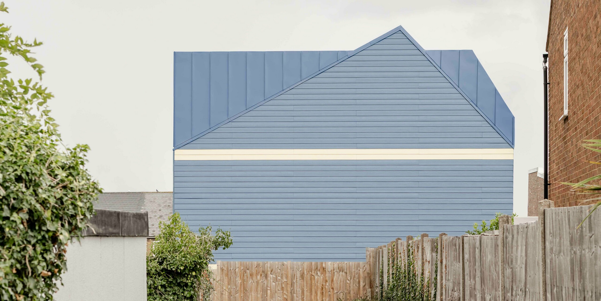 The photo shows the striking front of a Goldsmith Mews terraced house clad in dusty blue standing seam FALZONAL aluminium. The clean lines of the vertical and horizontal folded structures create a powerful, geometric façade that stands out clearly from the surrounding buildings. The contrast with the light-coloured lower façade areas reinforces the modern, precise design. The durable, colour-stable PREFA aluminium offers high weather protection and ensures a lasting high-quality appearance, giving the ensemble its memorable identity.