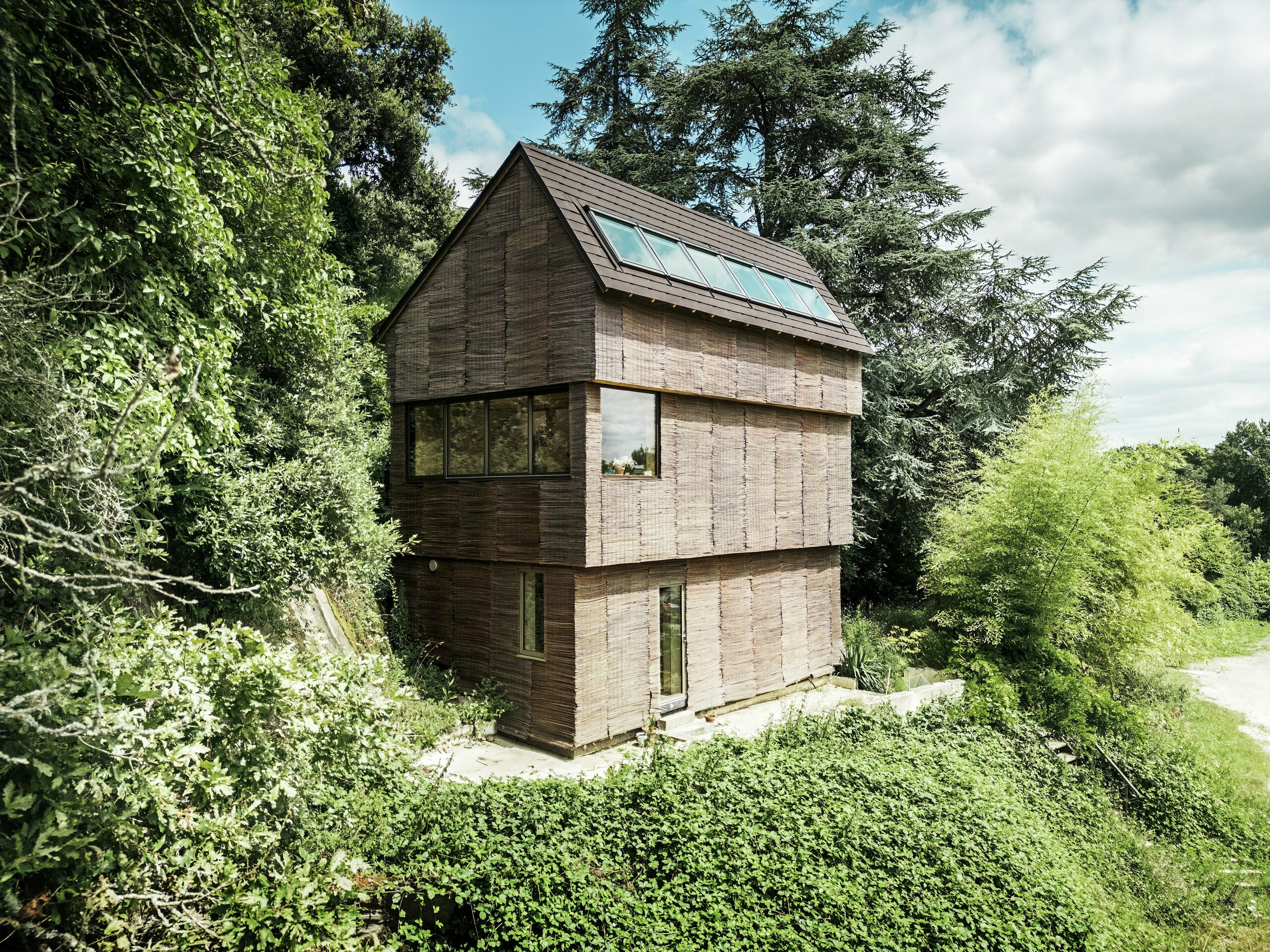 Ein Haus in Frankreich mit PREFA-Dachschindeln in der Farbe P.10 Walnussbraun und einer Fassade aus Weidenholz, umgeben von natürlichem Grün – nachhaltige Architektur und umweltfreundliches Design.