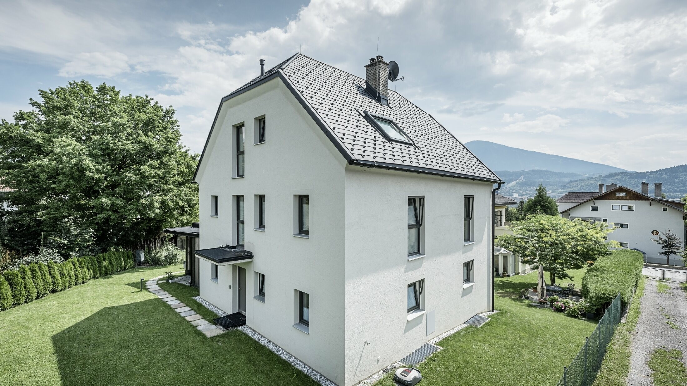 Modern three-storey detached house with a light-coloured plaster façade and dark grey aluminium gable roof from PREFA. The building has several narrow windows and anthracite-coloured roof drainage. It stands in a well-kept garden with a lawn, trees and hedges, with other residential buildings and a mountainous landscape visible in the background.