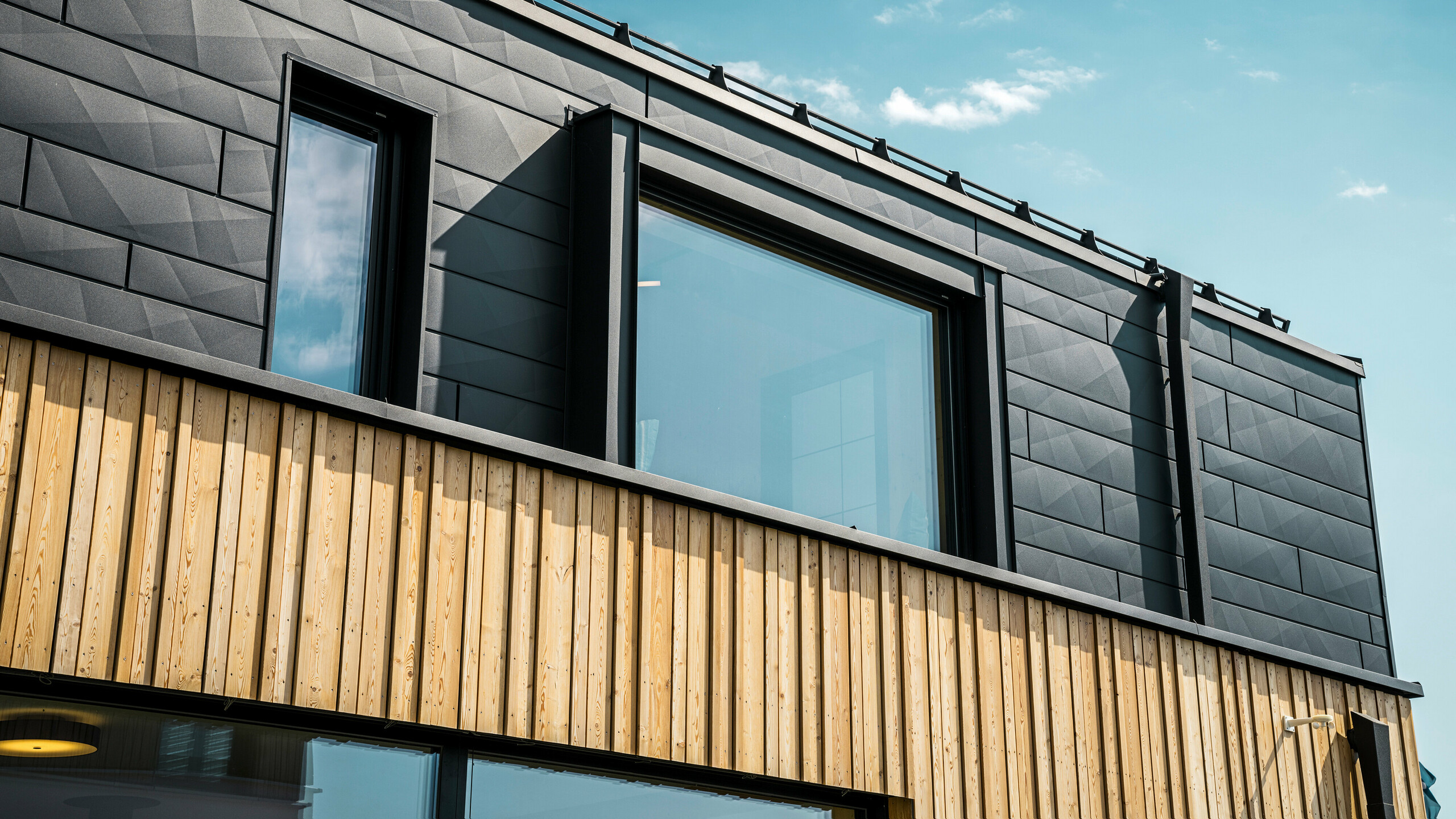 Nahaufnahme des Obergeschosses des Musterhauses Autark im Fertighauszentrum Blaue Lagune, Vösendorf (AT). Die Fassade besteht aus PREFA Siding.X Aluminiumpaneelen in P.10 Schwarz, die durch ihre spezielle Optik mit unterschiedlichen Kantungen auffallen. Darunter befindet sich eine stilvolle Holzverkleidung, die einen harmonischen Kontrast zum dunklen Aluminium bildet. Große Fenster lassen viel Licht in das Gebäudeinnere und betonen das moderne Design.