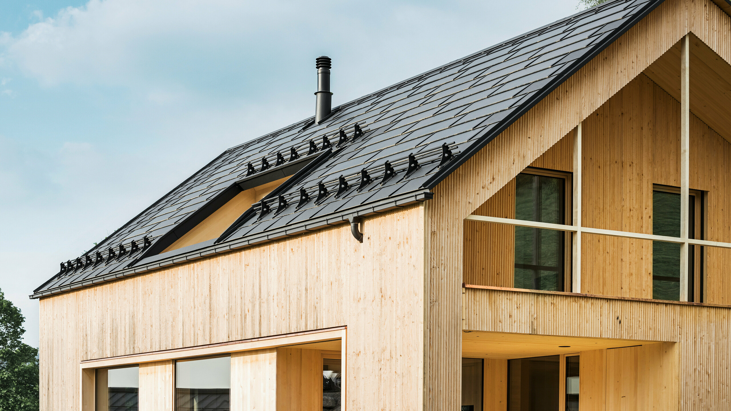 Close-up of the gable of an environmentally friendly house in Egg, characterised by PREFA solar roof tiles in elegant black, which blend harmoniously into the centre of the PREFA R.16 roof tiles. The clean lines of the roof contrast with the natural texture of the wooden façade, while the solar modules make a statement for sustainable living. The subtle chimney blends into the overall picture, which combines environmentally conscious design with rural aesthetics. In addition, the innovative eco-house has two balconies that offer a view of the beautiful mountain scenery of Egg in Vorarlberg.