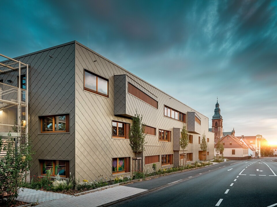 Im sanften Licht des Sonnenuntergangs zeigt sich die Kindertagesstätte in Alzenau von ihrer stimmungsvollsten Seite. Die Fassade aus bronzefarbenen PREFA Wandrauten 44 × 44 reflektiert das Abendlicht und betont die hochwertige, strukturierte Oberfläche des modernen Hybridgebäudes. Die klar gegliederte Architektur mit versetzten Fensterbändern und vertikalen Holzlamellen steht in spannendem Kontrast zur historischen Kirche im Hintergrund. Links im Bild sorgt ein filigranes Metallgerüst für leichte Transparenz beim Außentreppenhaus. Der gepflegte Grünstreifen entlang des Gebäudes unterstreicht den ökologischen Anspruch. Dieses Bild verbindet eindrucksvoll moderne Baukultur, nachhaltige PREFA Aluminiumtechnologie und harmonische Einbettung in das historische Stadtbild von Alzenau.
