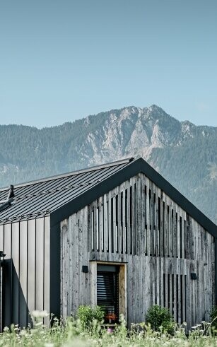 Moderner, funktionaler Wohnbau mit einer Kombination aus Holzfassade und PREFA Band- und Blechmaterial für das Dach. Die klare Architektur wird durch die elegante, metallische Dach- und Fassadenverkleidung unterstrichen, die ideal für anspruchsvolle Projekte ist. Das Bild veranschaulicht die vielseitigen Anwendungsmöglichkeiten der PREFA Produkte im Bereich Bänder und Bleche.