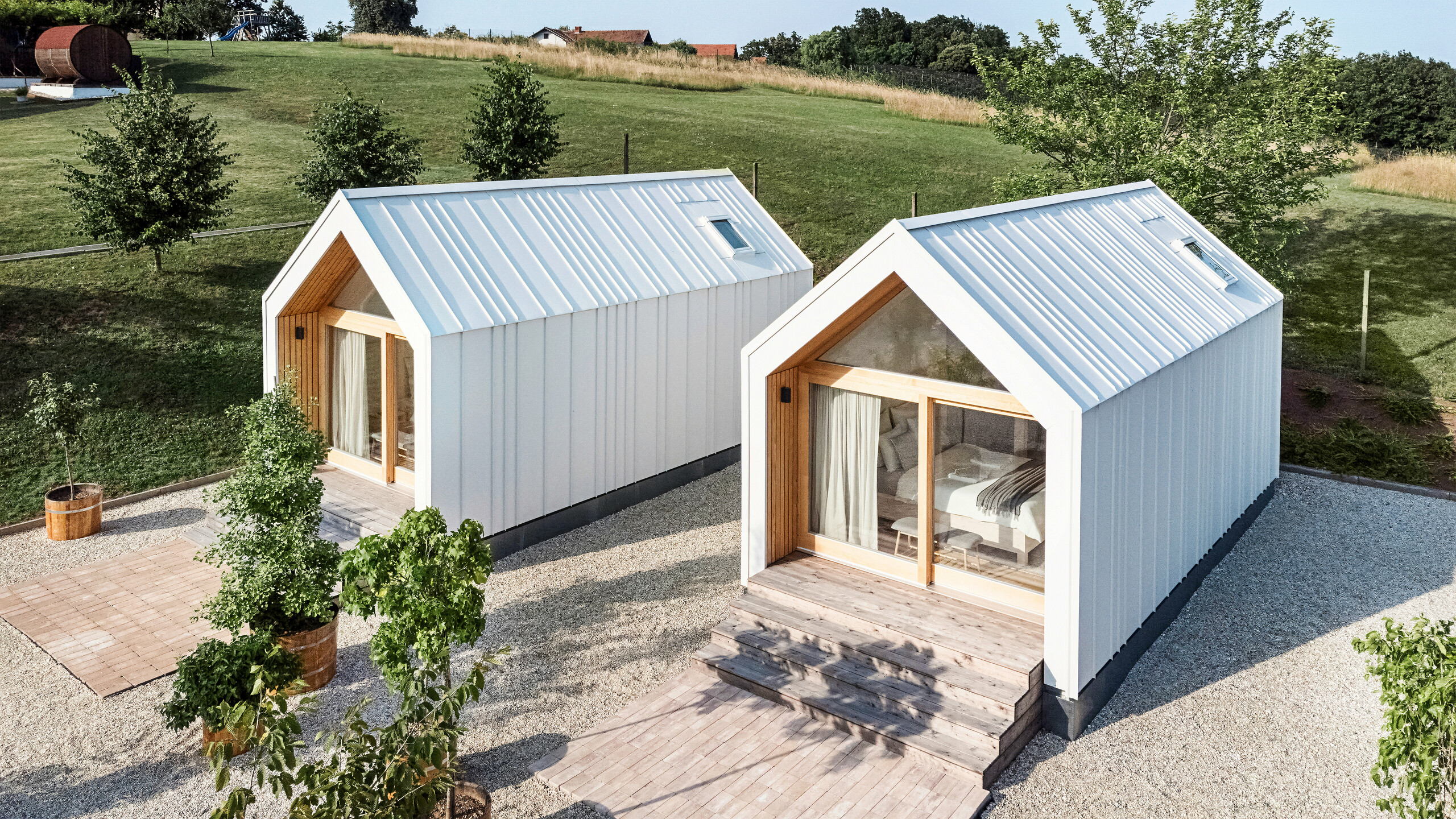 View of the front of the two tiny houses in the 'Pri Momi' holiday complex in Kančevci, Slovenia. The micro houses were built using a timber frame construction and finished with a robust PREFALZ standing seam roof and façade cladding in P.10 Pure White. The modern, minimalist buildings combine a durable yet aesthetic aluminium shell with large glass fronts and natural wood, embedded in a green landscape. The generous wooden steps lead up to the entrances, creating a cosy and inviting atmosphere.