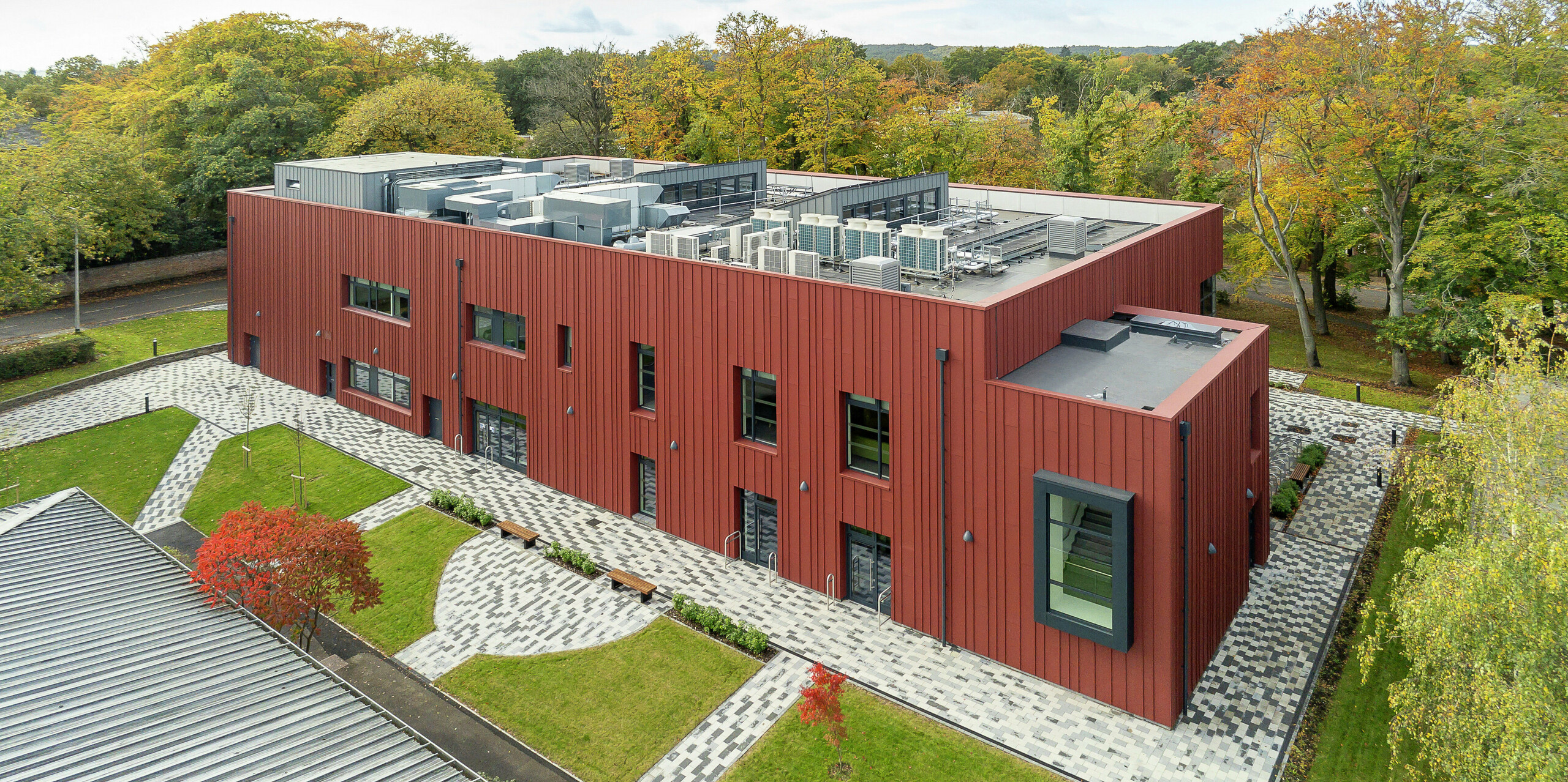 Architecture in harmony with nature: a bird's eye view of the Soldiers' Centre in Pirbright. The distinctive standing seam façade made of PREFALZ in P.10 oxide red blends harmoniously into the green surroundings. The cantilevered window frames in P.10 anthracite set modern accents. The structured outdoor area with paved paths, green spaces and seating creates an inviting atmosphere. Technical installations are integrated on the flat roof, contributing to the modern building technology.