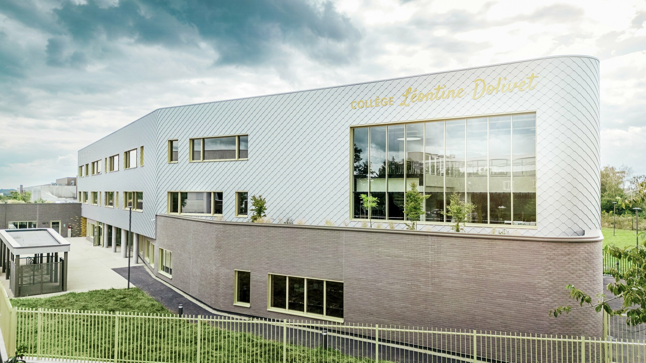 Seitenansicht des Collège Léontine Dolivet in Cesson-Sévigné mit seiner modernen Aluminiumfassade aus PREFA-Wandelementen in Silbermetallic. Die diagonale Struktur verleiht dem Gebäude eine dynamische Optik, während die großen Glasflächen und gelben Fensterrahmen interessante Akzente setzen. Im Vordergrund sind die Grünflächen und der das Gebäude umgebende Zaun zu sehen. Die langlebige PREFA-Fassadenlösung vereint Eleganz, Witterungsbeständigkeit und Nachhaltigkeit – ein architektonisch ansprechendes Gebäude mit klaren Linien und variablem Metall.