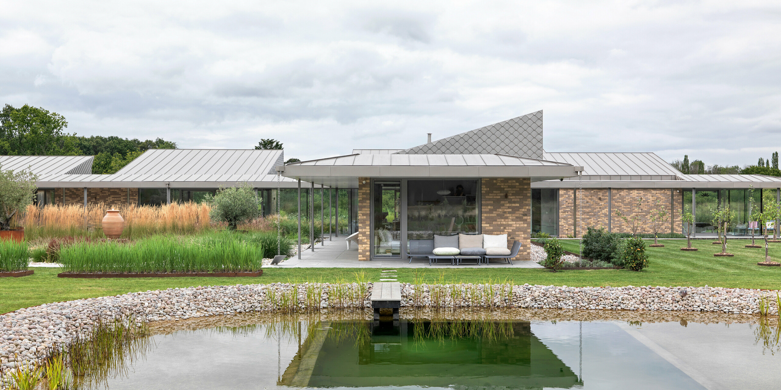 Symmetrical overall view of the residential building in West Sussex with standing seam roof in P.10 bronze and façade accents with PREFA rhomboid façade tiles 20 × 20. In the foreground is a landscaped natural pool with a wooden walkway, surrounded by a natural garden landscape.The clear architecture with large glazed areas and durable PREFA materials creates a modern, weather-resistant and aesthetically high-quality building ensemble.
