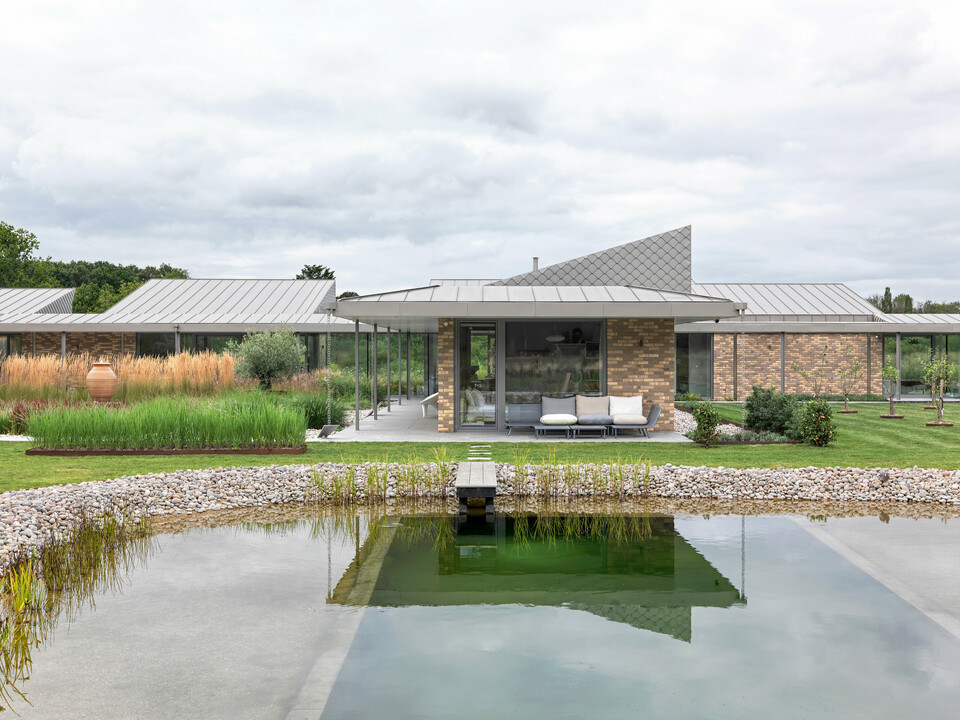 Symmetrical overall view of the residential building in West Sussex with standing seam roof in P.10 bronze and façade accents with PREFA rhomboid façade tiles 20 × 20. In the foreground is a landscaped natural pool with a wooden walkway, surrounded by a natural garden landscape.The clear architecture with large glazed areas and durable PREFA materials creates a modern, weather-resistant and aesthetically high-quality building ensemble.
