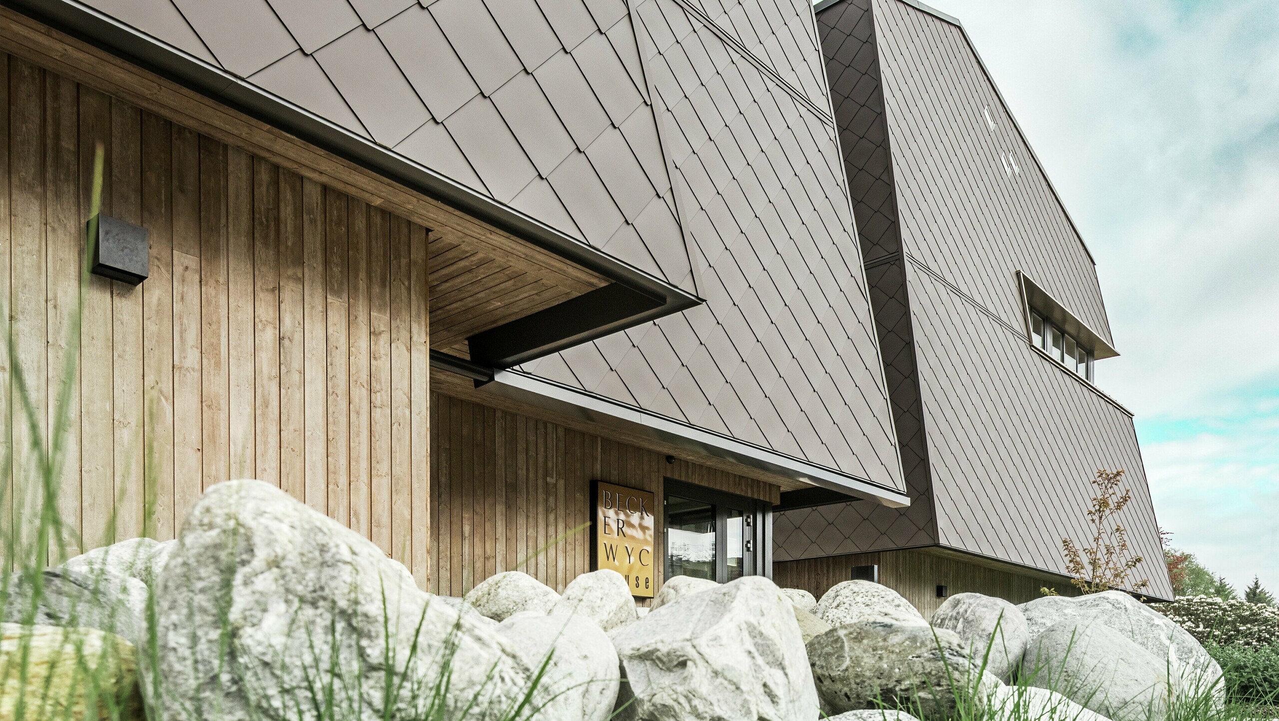 Side view of a hotel, the façade of the ground floor is vertically clad with rather dark wood. Above it is a façade projection, from here the façade is clad with aluminium panels in nut brown.